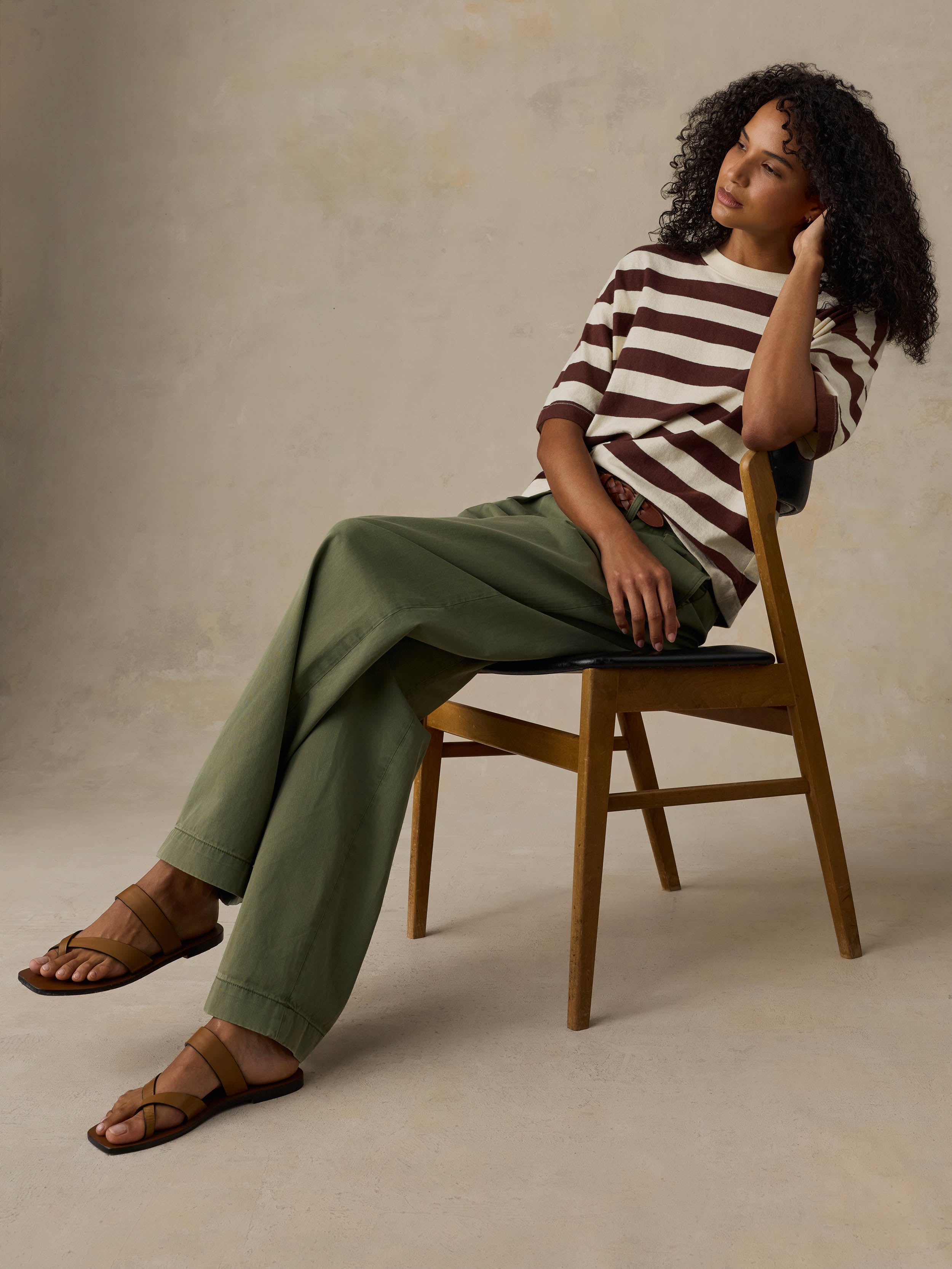 Woman sitting on a wooden chair wearing a brown and white striped shirt with green pants against a beige background