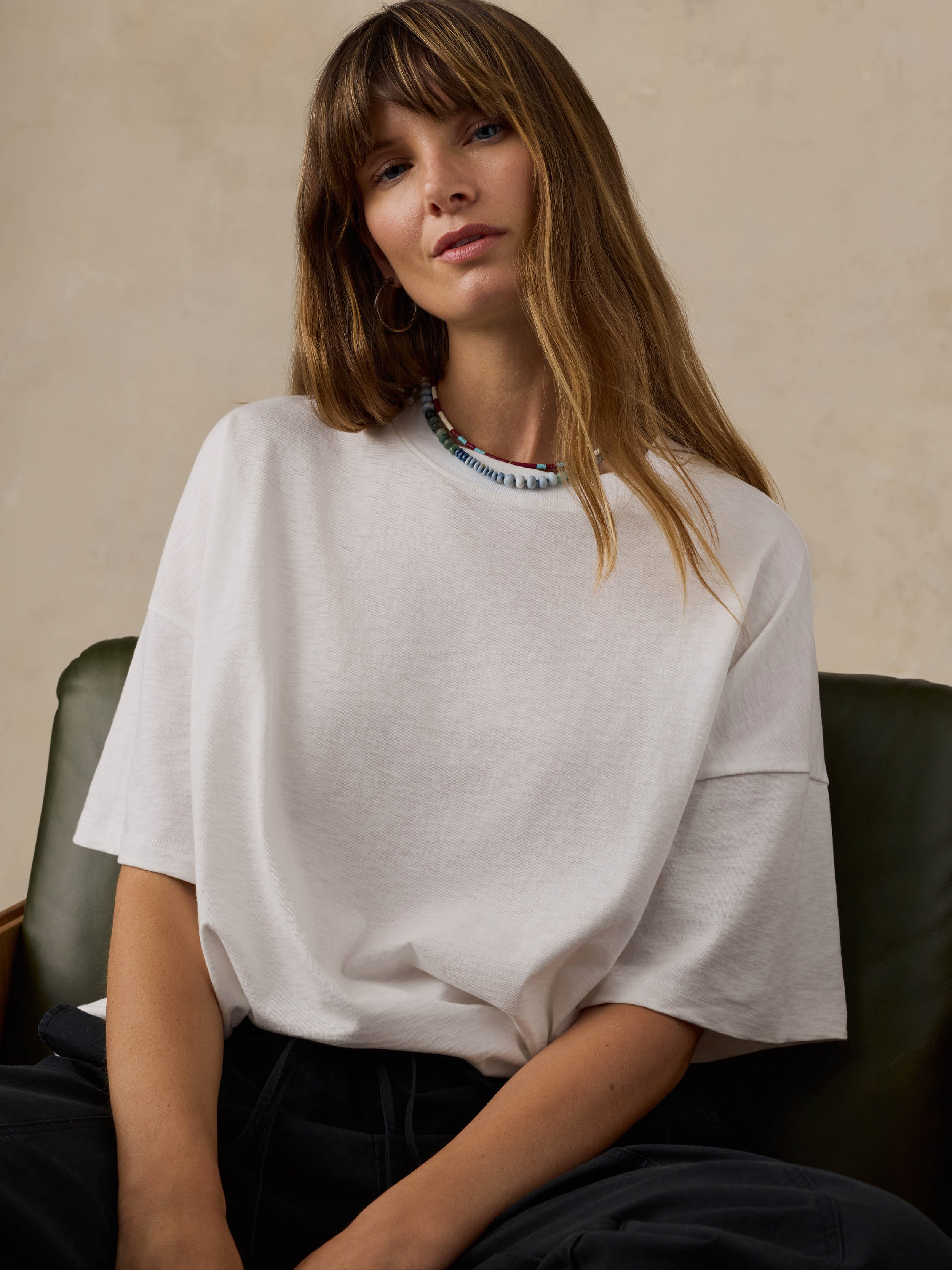 Woman sitting on a wooden chair wearing a white t-shirt and black pants against a beige background
