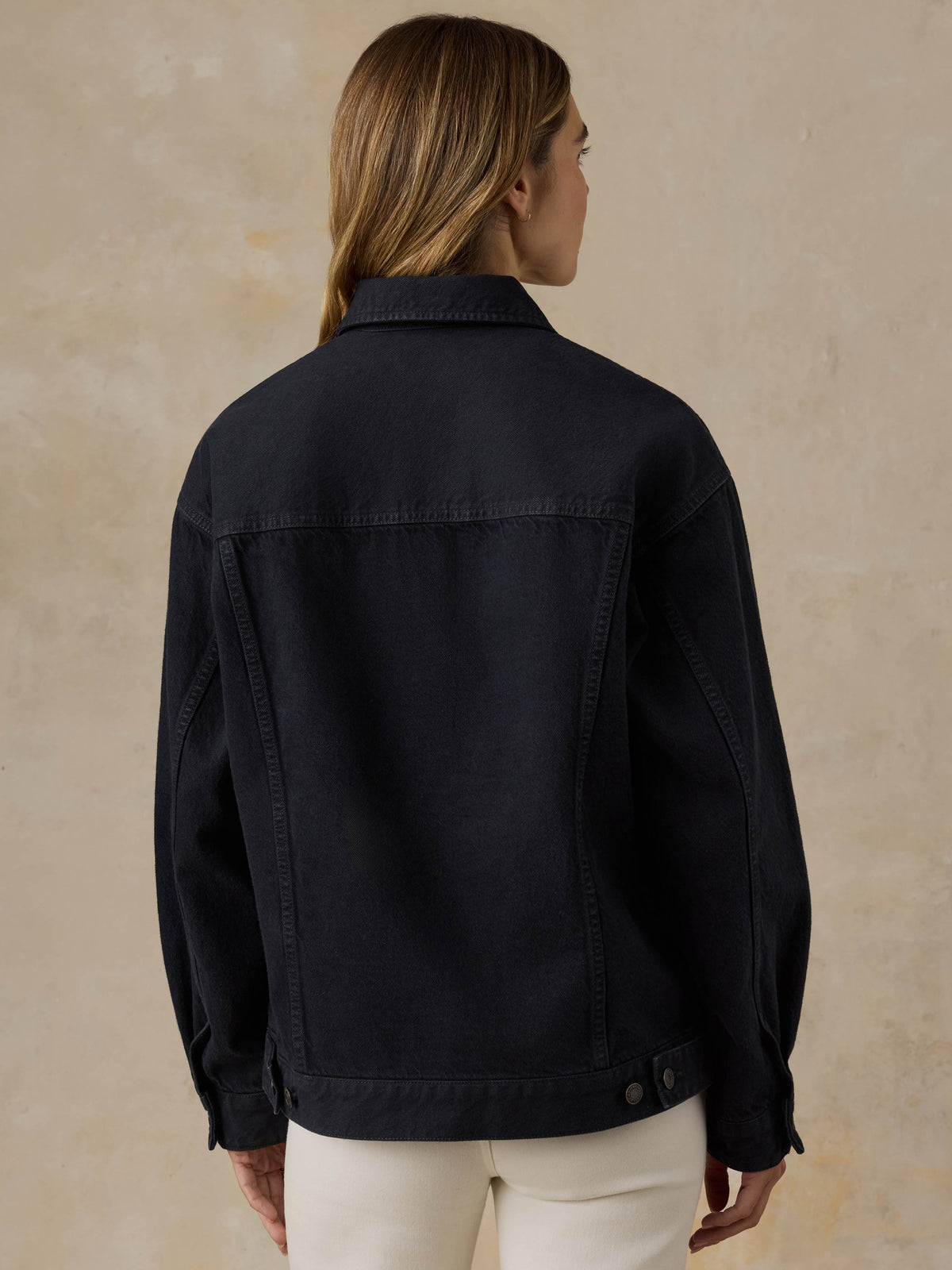 Woman wearing a black denim jacket and white jeans against a beige background, image 5 of 5