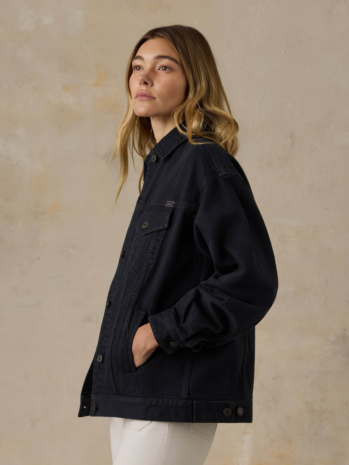 Woman wearing a black denim jacket and white jeans against a beige background, image 3 of 5
