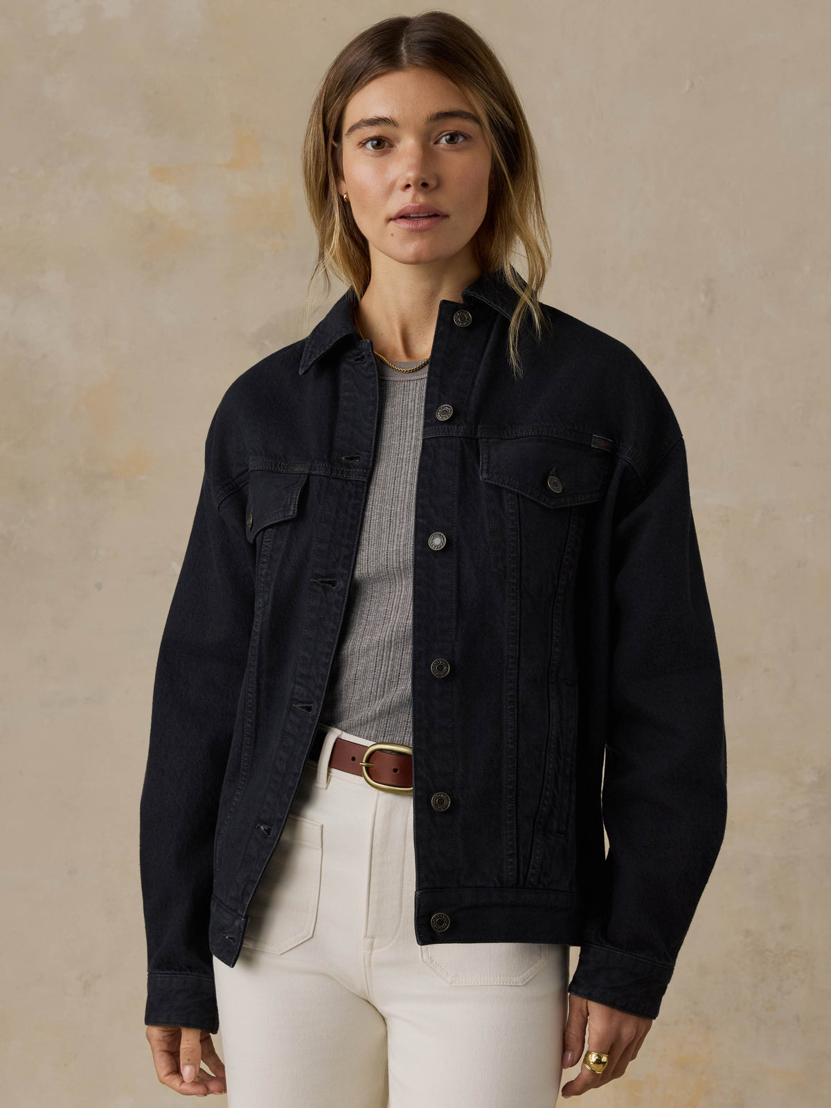 Woman wearing a black denim jacket and white jeans against a beige background, image 1 of 5