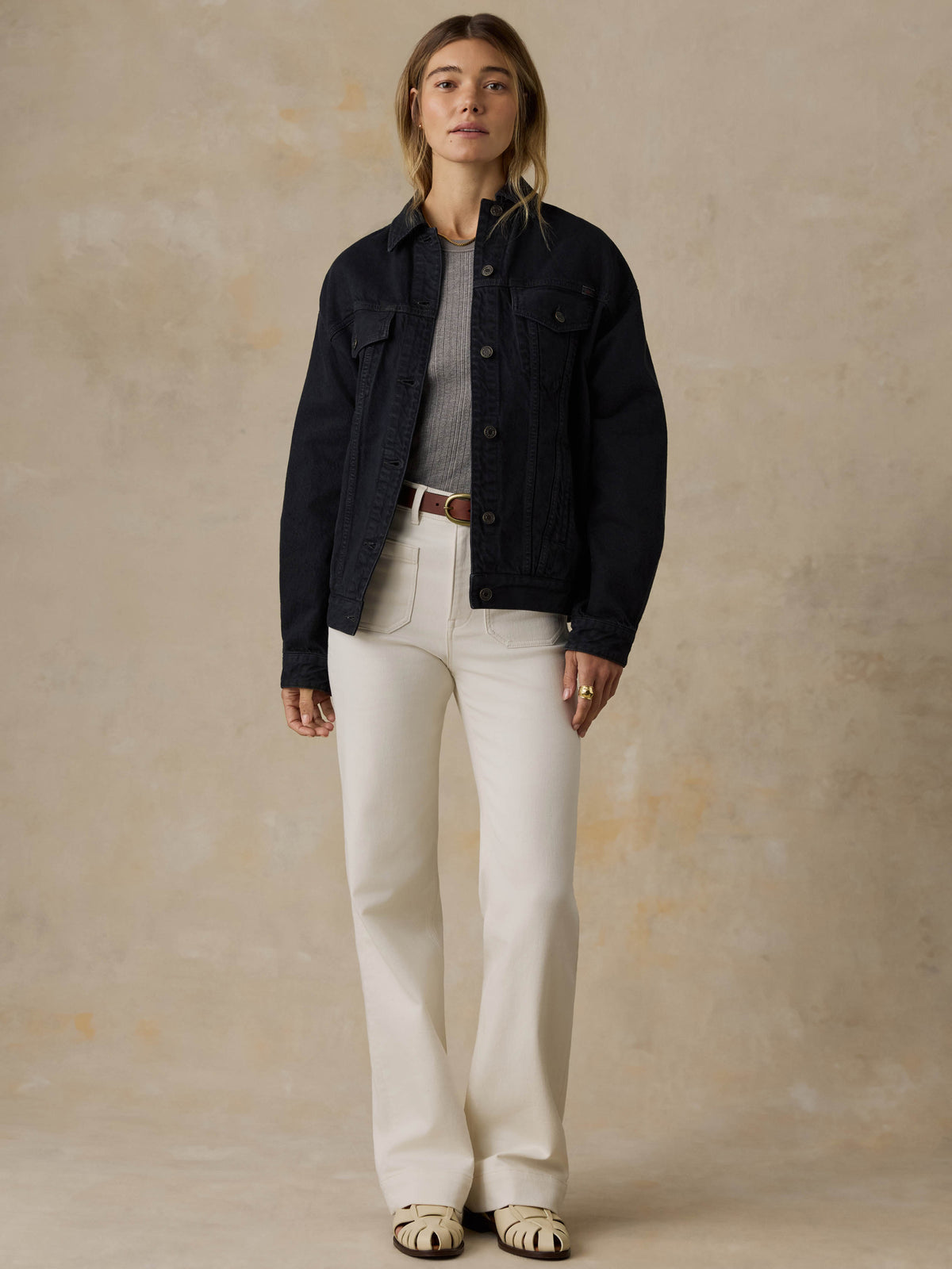 Woman wearing a black denim jacket and white jeans against a beige background, image 2 of 5
