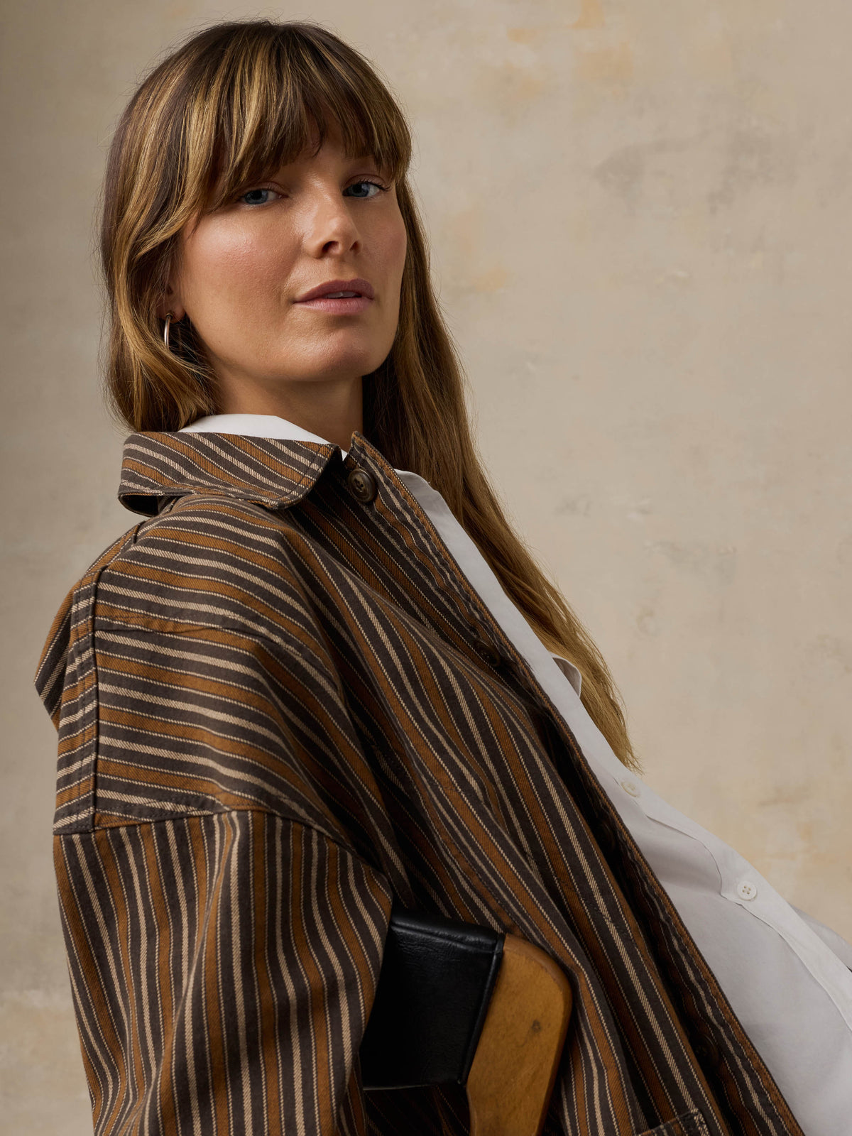 Woman sitting on a wooden chair wearing a brown striped chore jacket against a beige background, image 2 of 6