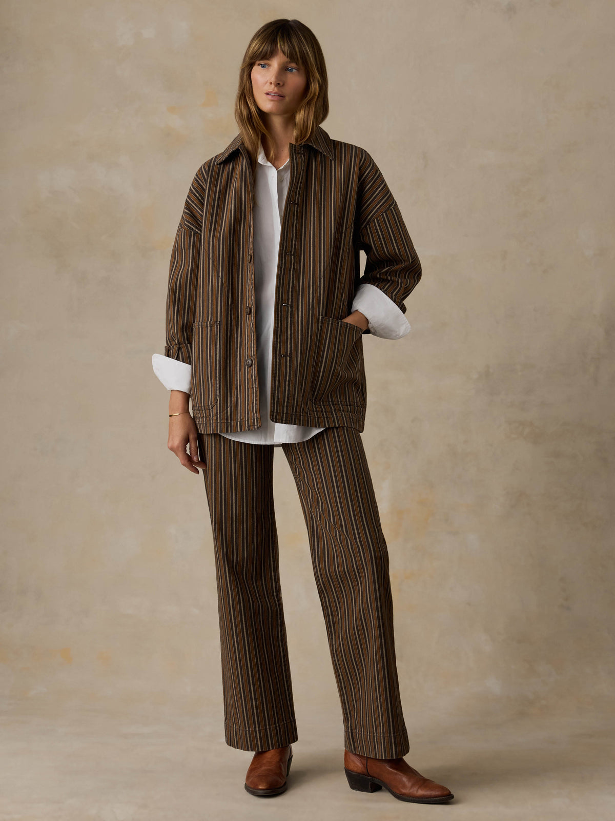 Woman wearing a white button down shirt, brown striped chore jacket, and matching striped pants against a beige background, image 6 of 6