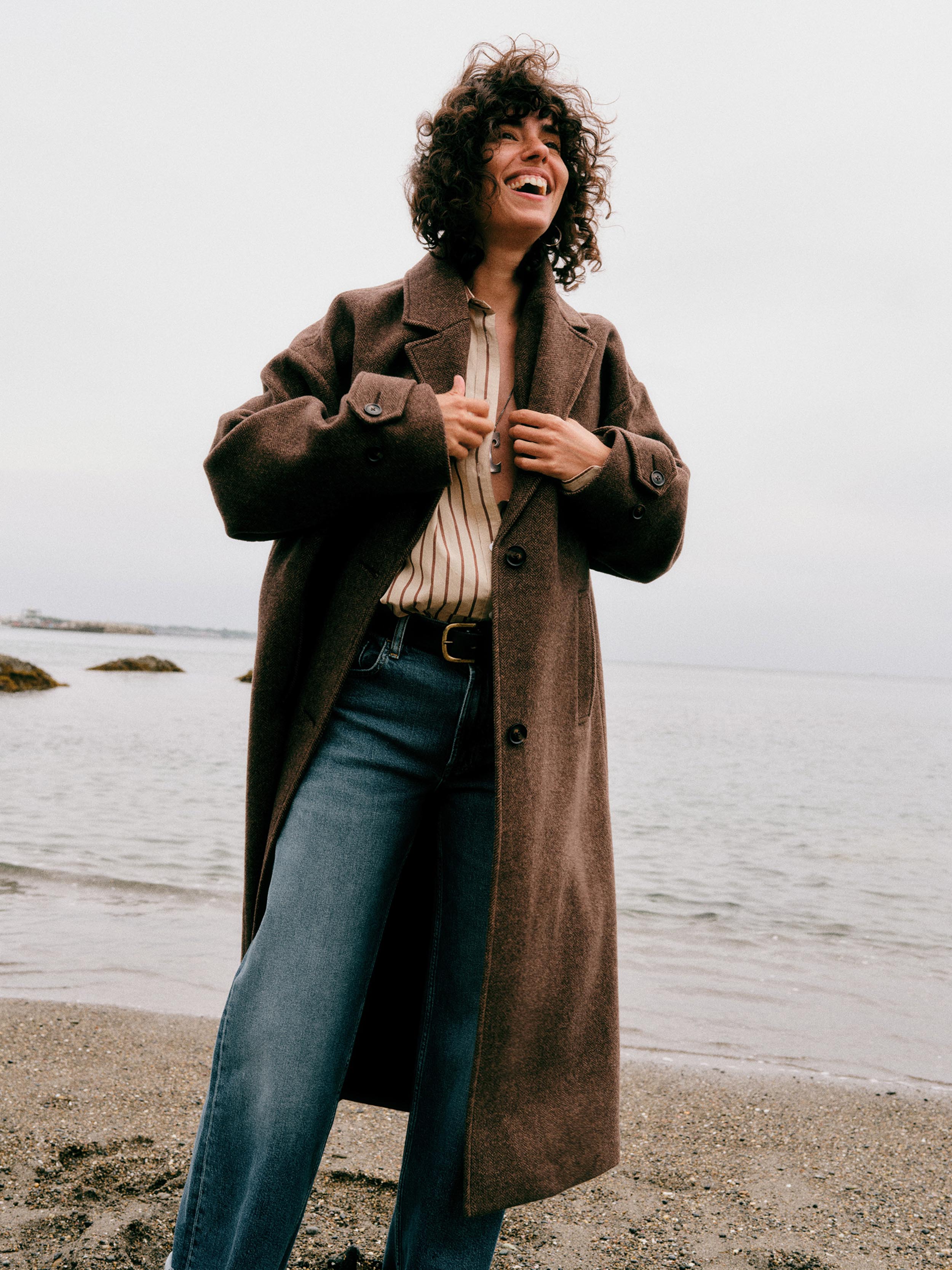 A woman wearing a brown coat over a striped shirt and blue jeans