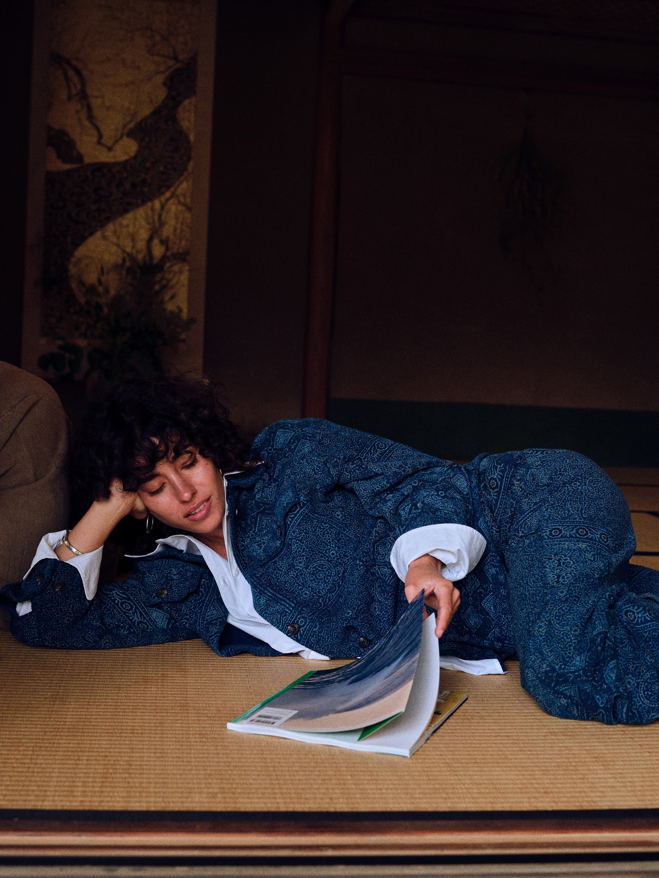 A woman reclines on a tatami mat floor wearing a dark blue patterned kimono jacket over a white collared shirt, holding an open magazine in a dimly lit interior setting.