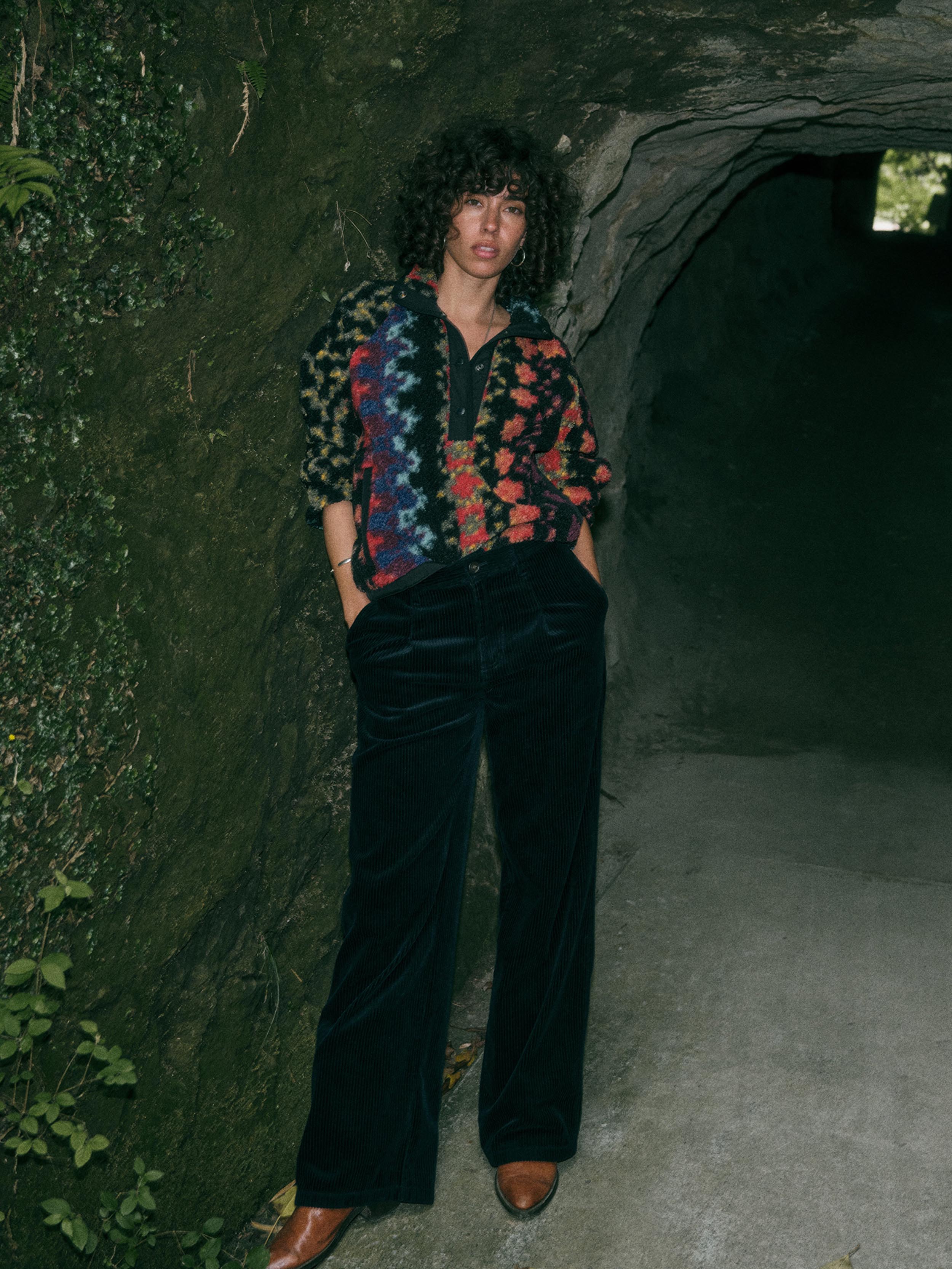 A model stands in a dark, mossy stone tunnel wearing a colorful patterned button-up shirt, black velvet wide-leg trousers, and brown leather shoes.