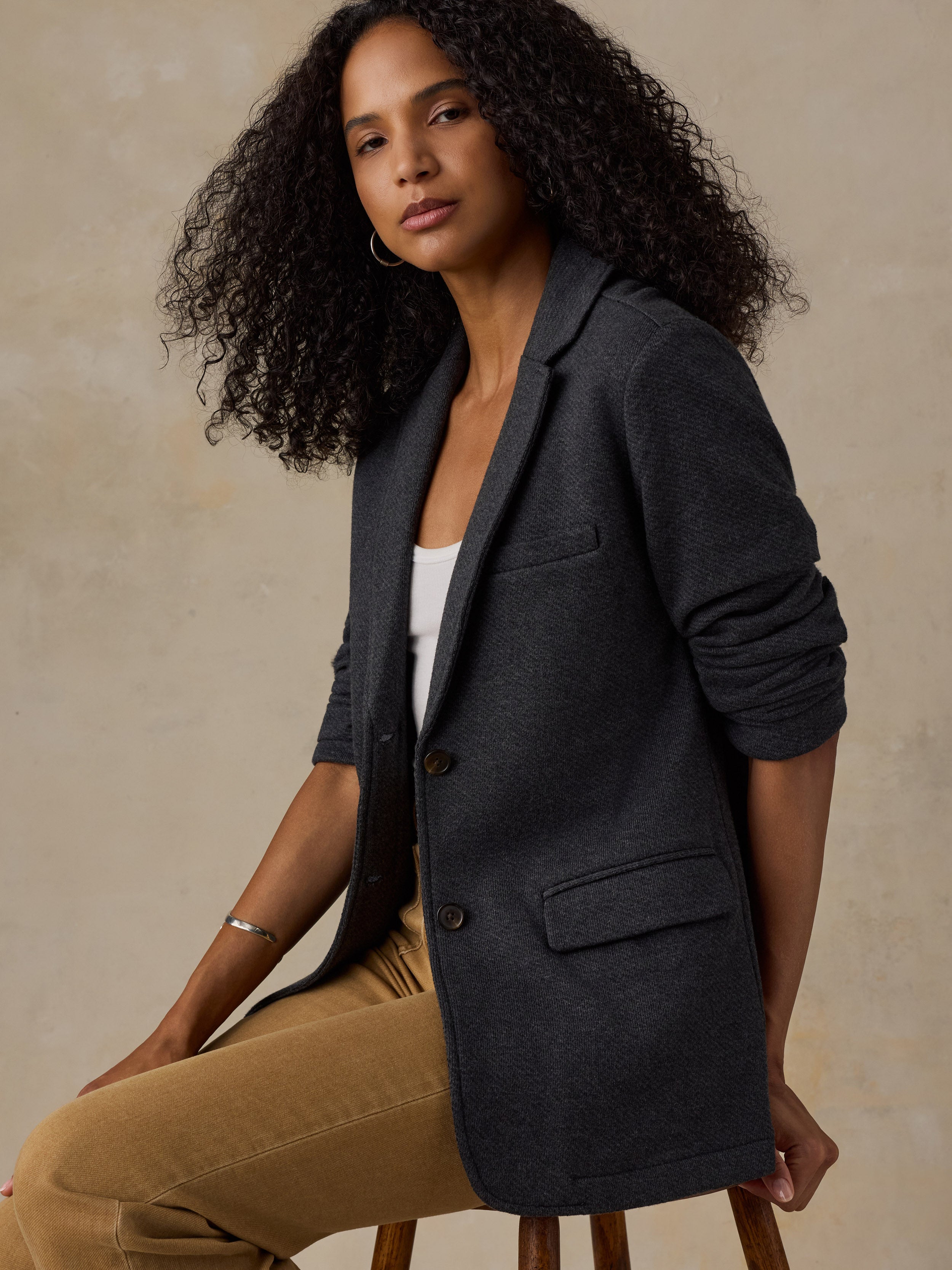 Woman sitting on a stool wearing a black blazer over a white tank and brown pants against a beige background