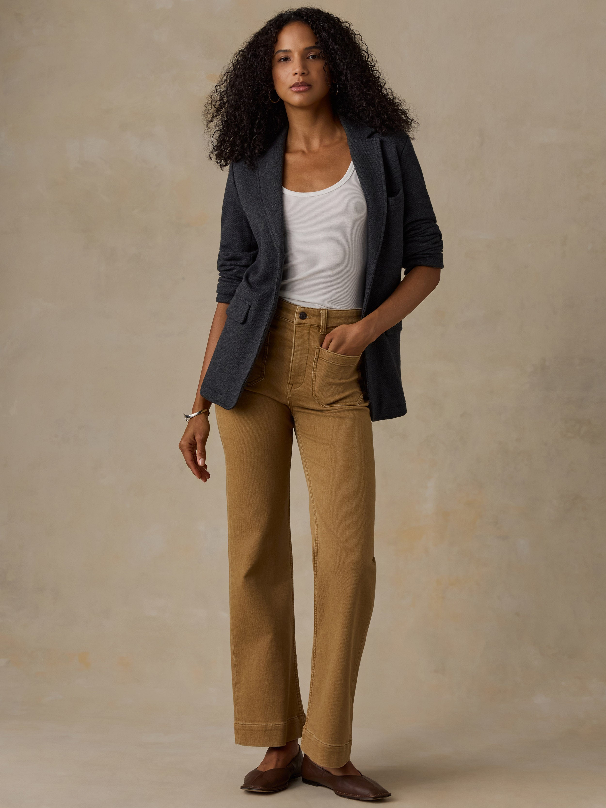 Woman wearing a black blazer over a white tank and brown pants against a beige background