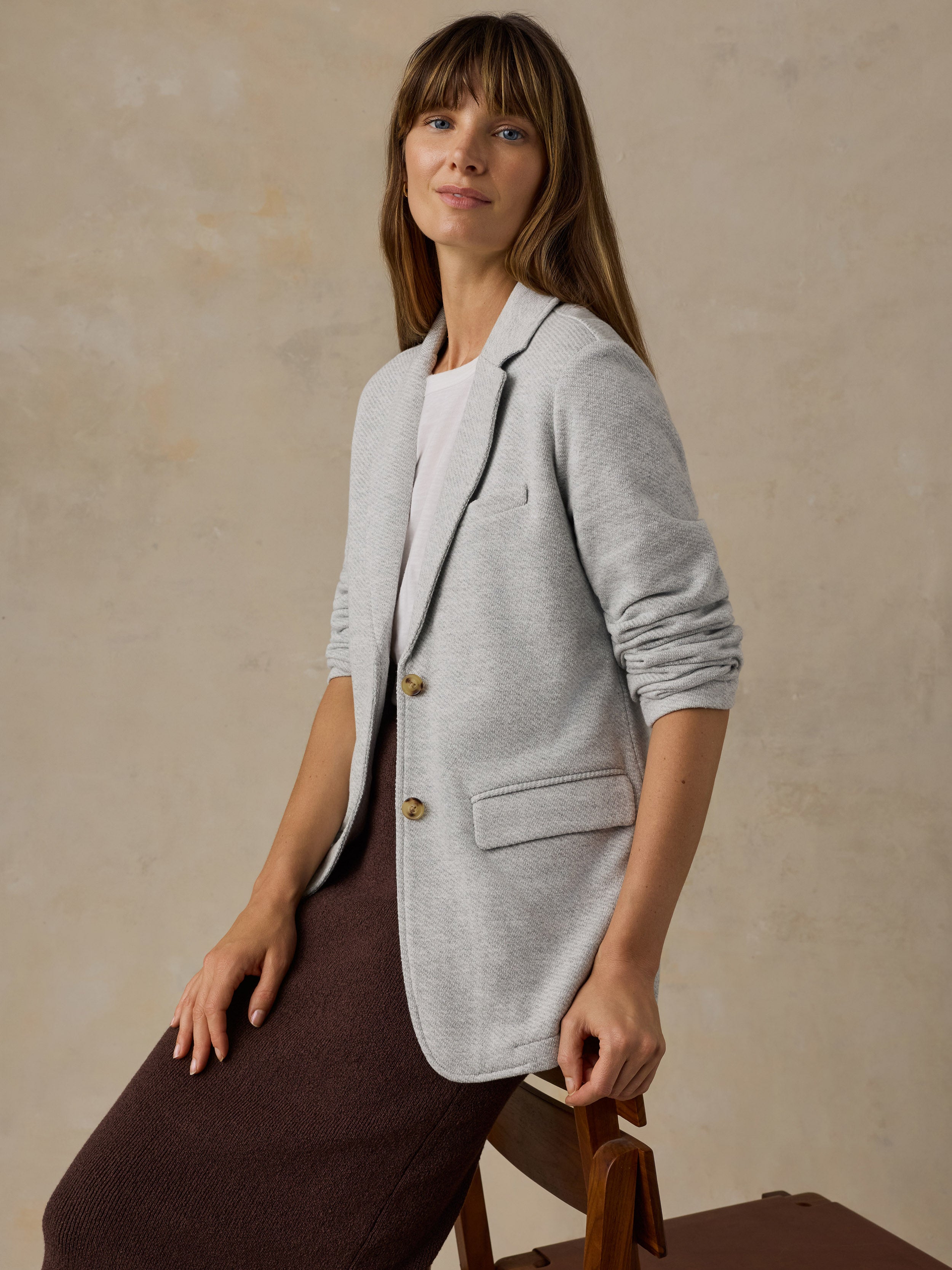 Woman sitting on a wooden chair wearing a grey blazer over a white t-shirt and brown skirt against a beige background
