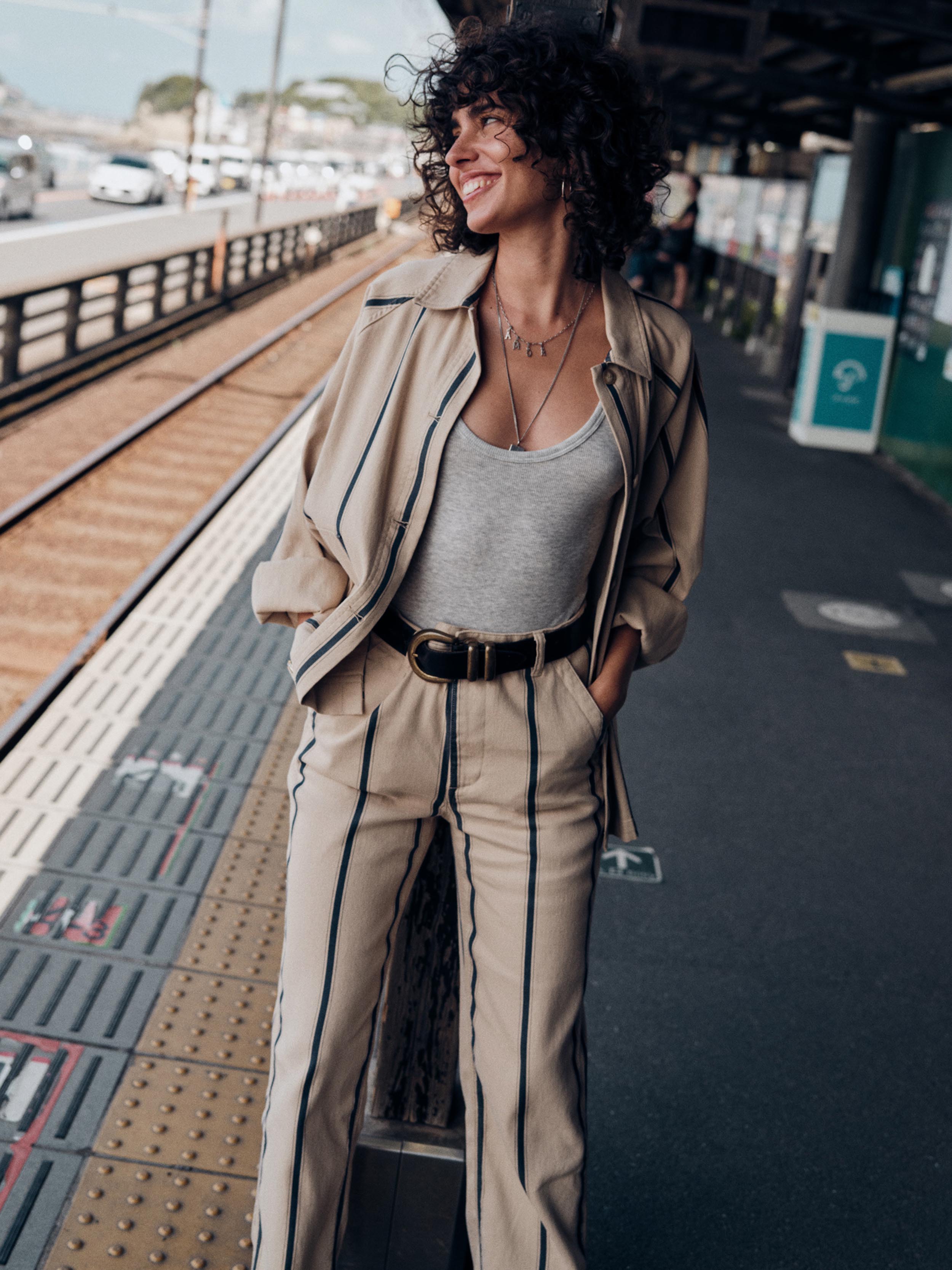 A woman with curly dark hair stands on a train station platform wearing a beige tailored suit with thin dark stripes, a light gray tank top, and a black belt.