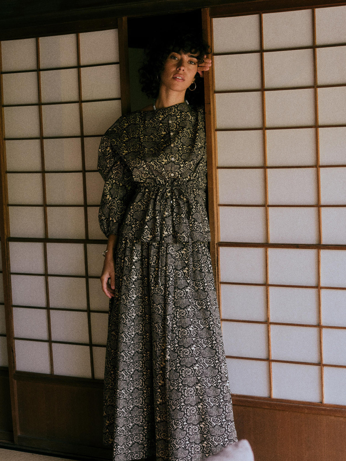 A woman wearing a long-sleeved, floor-length black and gray patterned dress stands in a doorway with traditional Japanese shoji screens behind her., image 1 of 7