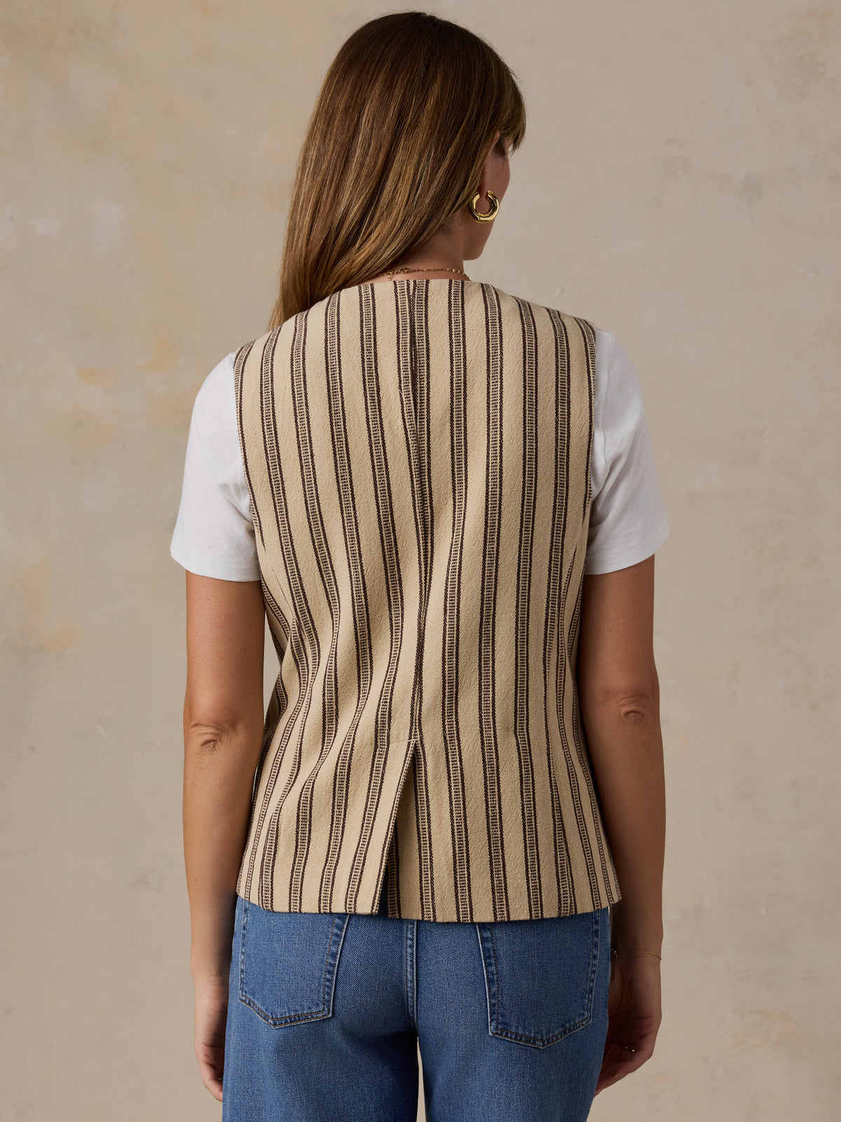 A woman viewed from the back wearing a sleeveless beige vest with vertical dark brown stripes over a white t-shirt and blue jeans, set against a textured beige background., image 3 of 5