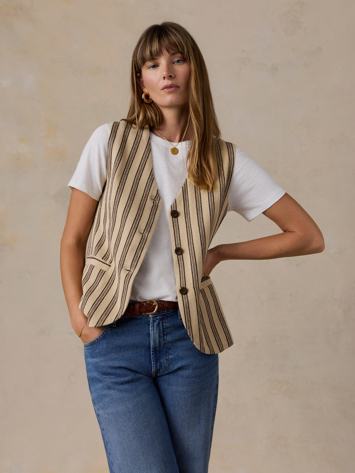 A woman modeling a beige and brown vertically striped button-up blazer vest over a white t-shirt and blue jeans, standing against a textured beige wall., image 5 of 5