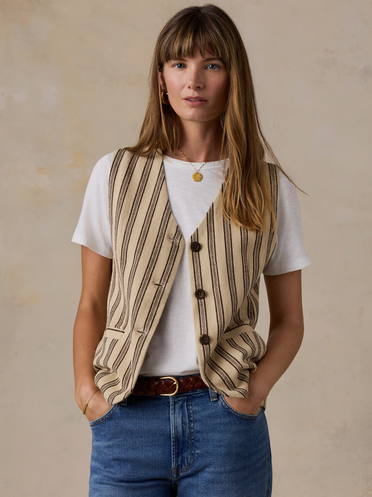 A woman wearing a beige and brown vertically striped knit vest over a white t-shirt, paired with blue jeans and a brown belt, standing against a textured beige wall., image 1 of 5