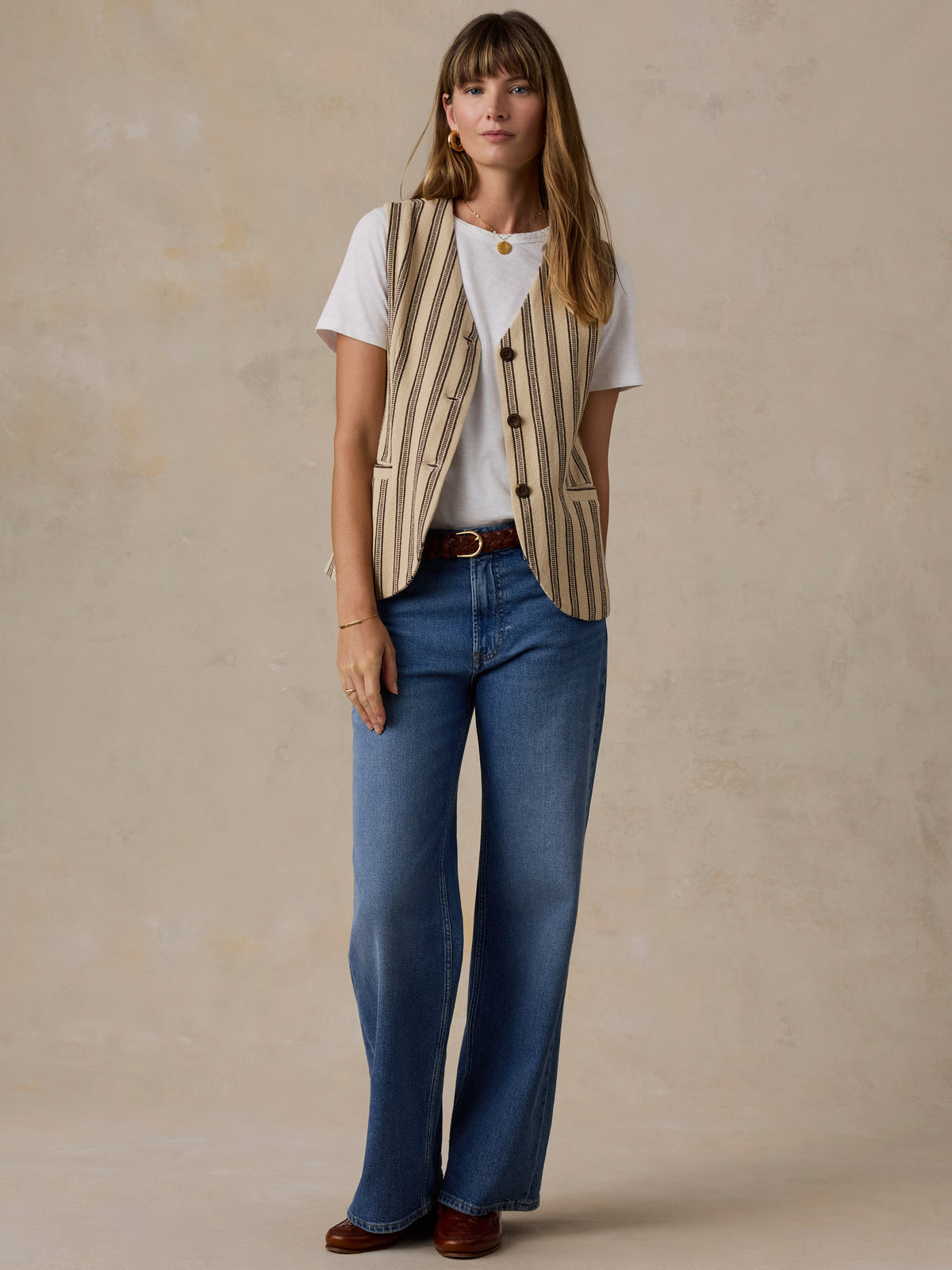 A woman stands against a textured beige studio backdrop wearing a cream and tan vertically striped vest over a white T-shirt, blue jeans, and brown shoes., image 2 of 5