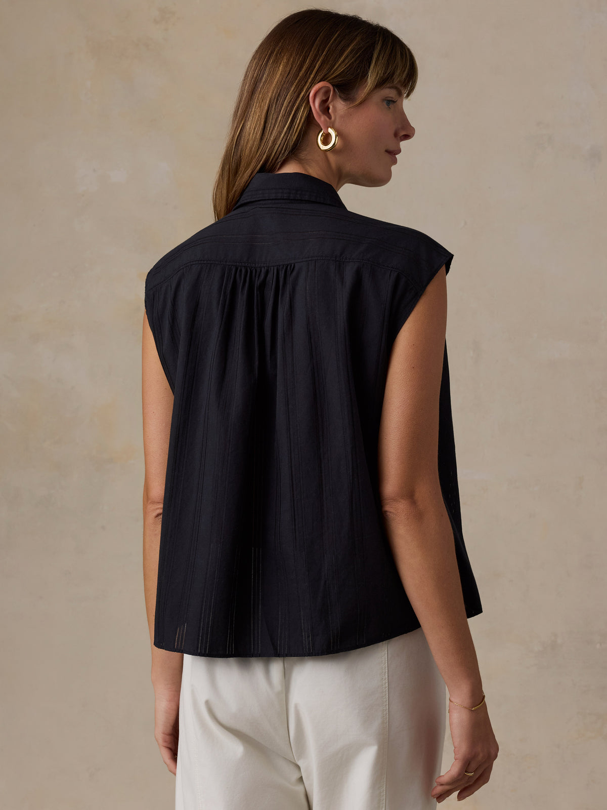 A woman shown from the back wearing a sleeveless black top with a gathered neckline and white pants, standing against a textured beige wall., image 3 of 5