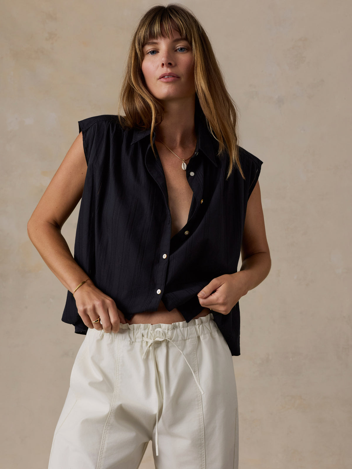 A woman wearing a sleeveless black button-up shirt and cream drawstring trousers, standing against a textured beige wall., image 2 of 5