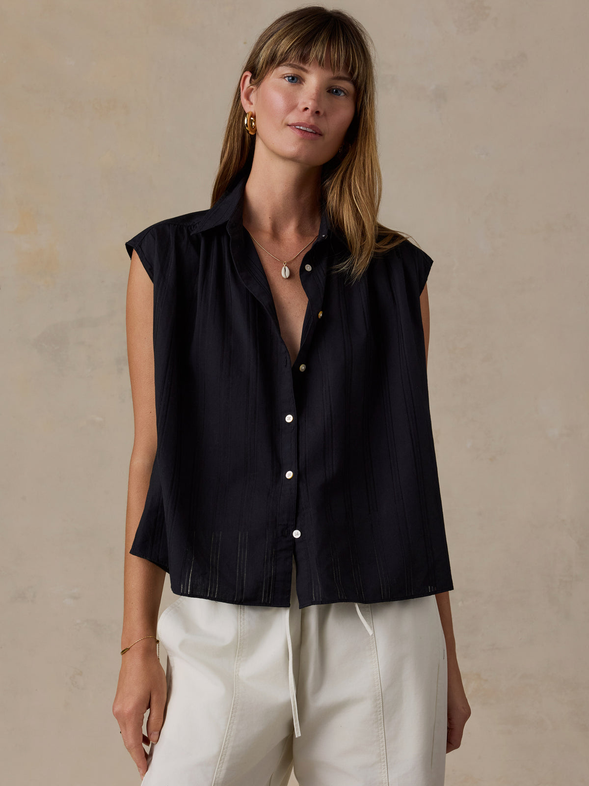 A woman wearing a sleeveless black button-front shirt with white drawstring pants, standing against a textured beige wall., image 1 of 5
