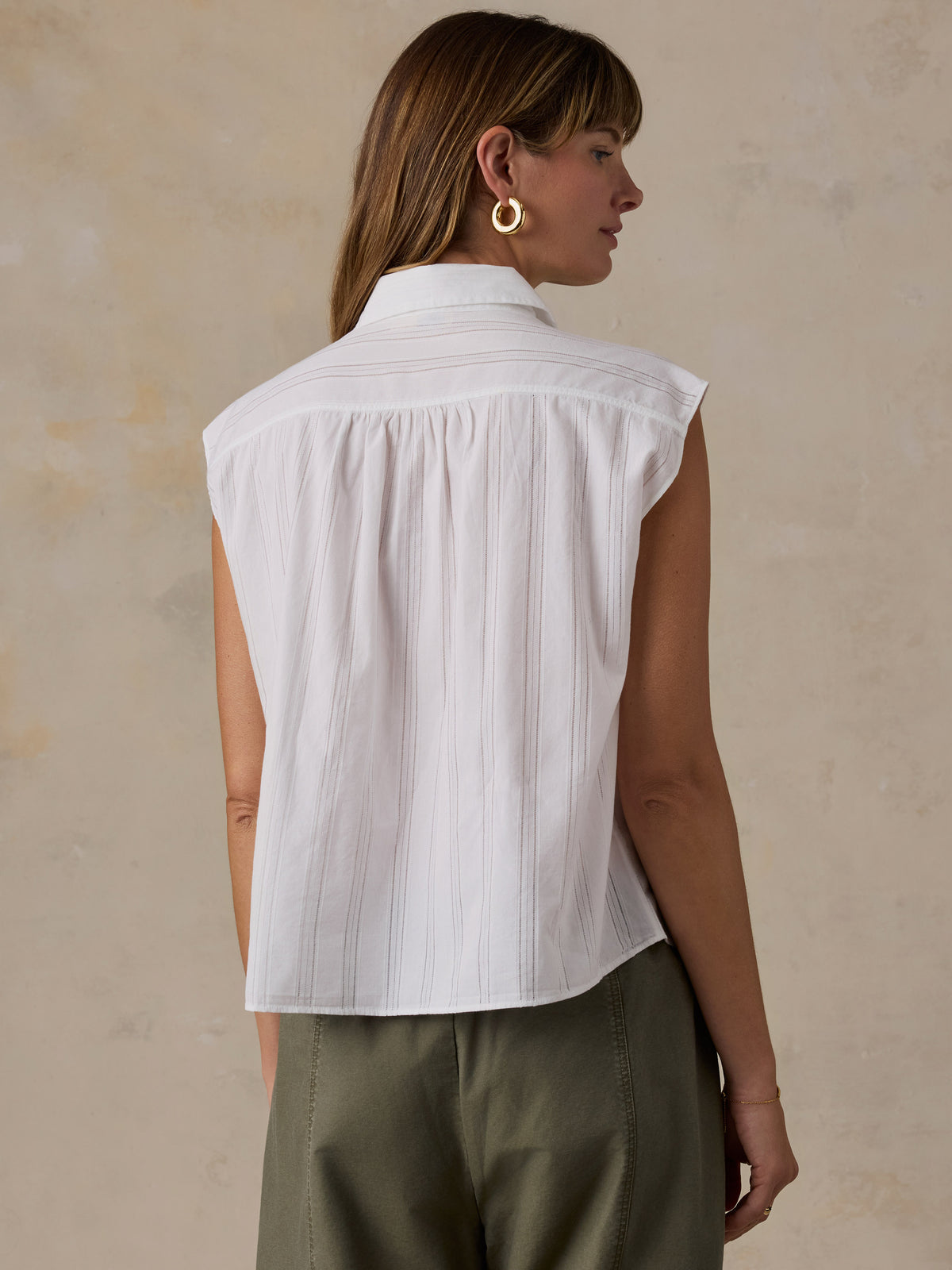 A woman shown from the back wearing a white sleeveless collared blouse with subtle vertical texture and pleated detailing, paired with olive green pants, against a textured beige wall., image 4 of 5