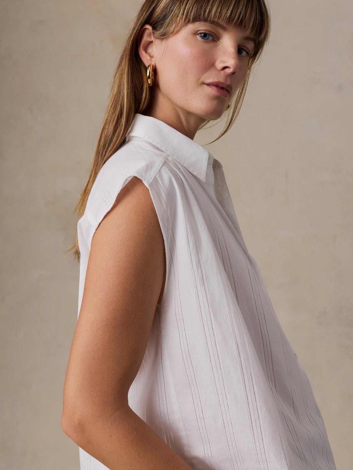 A woman wearing a sleeveless white collared blouse with a subtle vertical pleated texture, photographed against a warm beige textured background., image 3 of 5
