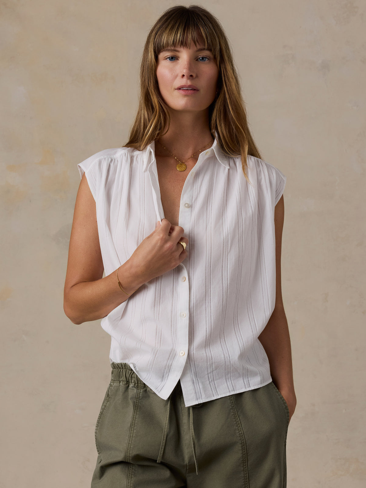 A woman wearing a white sleeveless button-up shirt with subtle vertical pinstripes and olive green pants, standing against a textured beige studio background., image 1 of 5