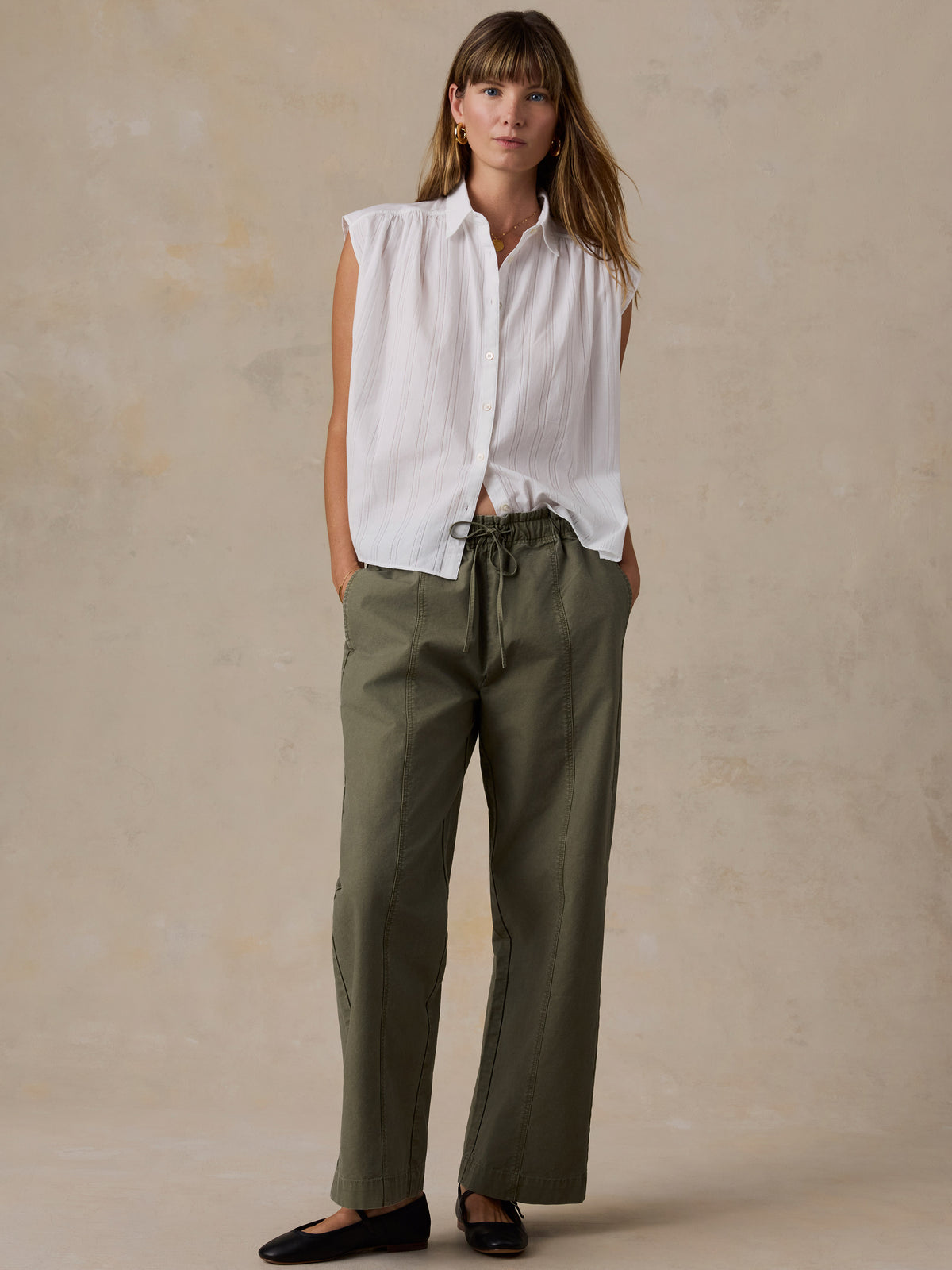 A woman stands against a textured beige studio backdrop wearing a sleeveless white button-up shirt and olive green drawstring pants with black flats., image 2 of 5