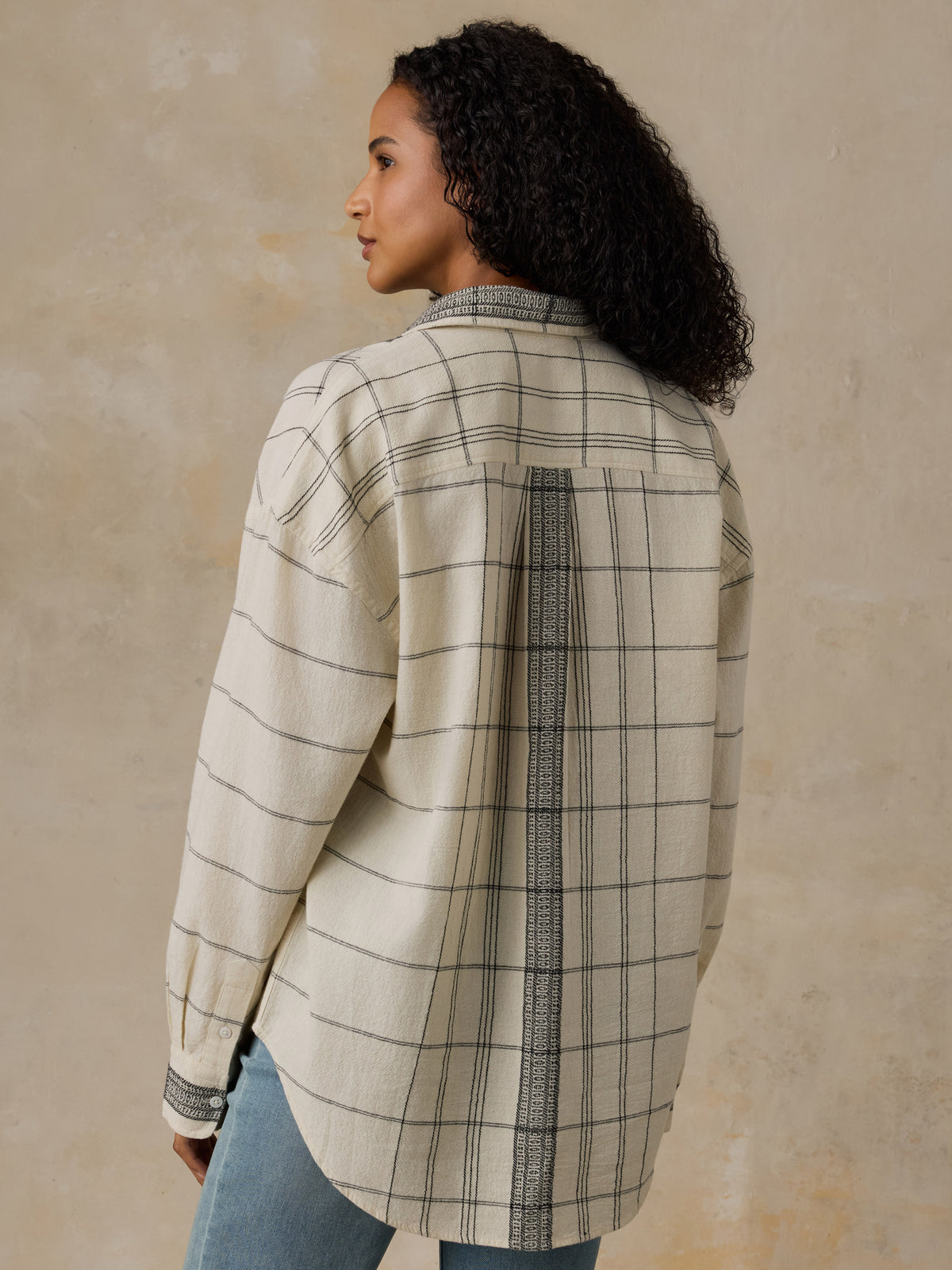 Woman wearing a white and black plaid shirt with blue jeans against a beige background, image 3 of 6