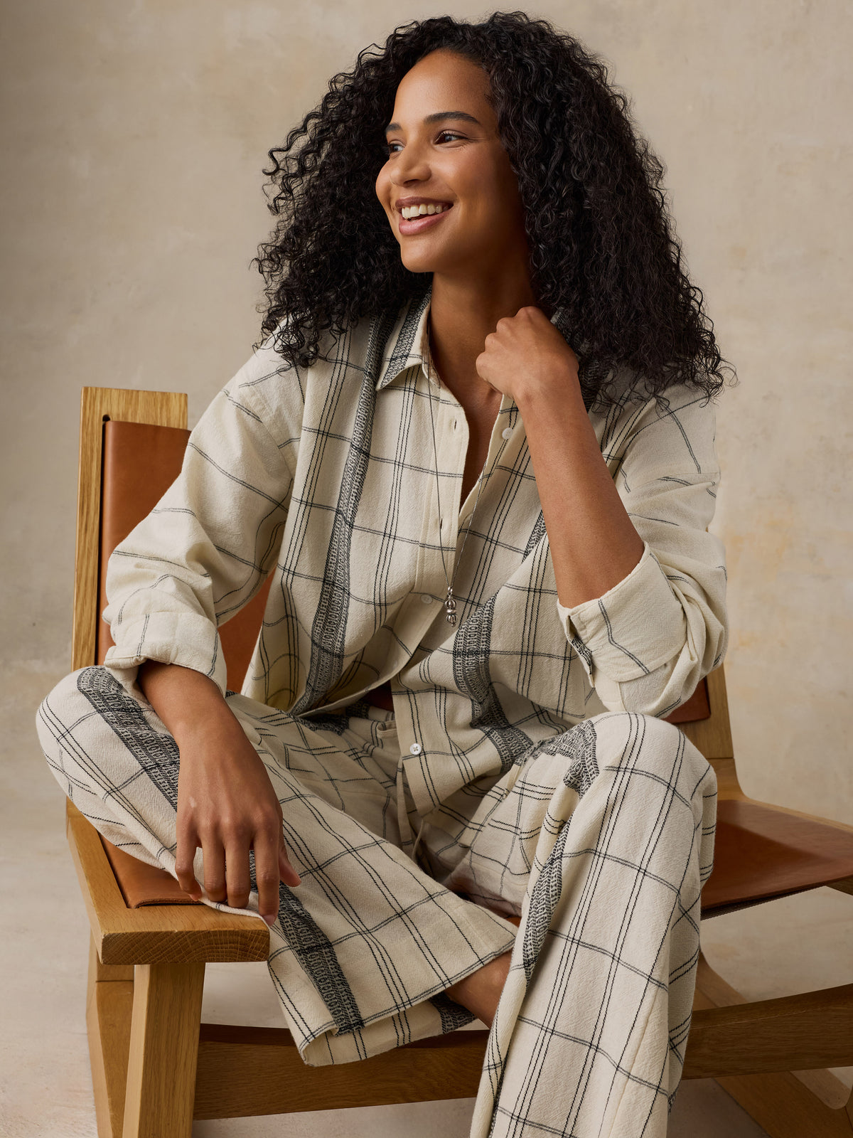 Woman sitting on a wooden chair wearing a white and black plaid shirt and matching pants against a beige background, image 2 of 6