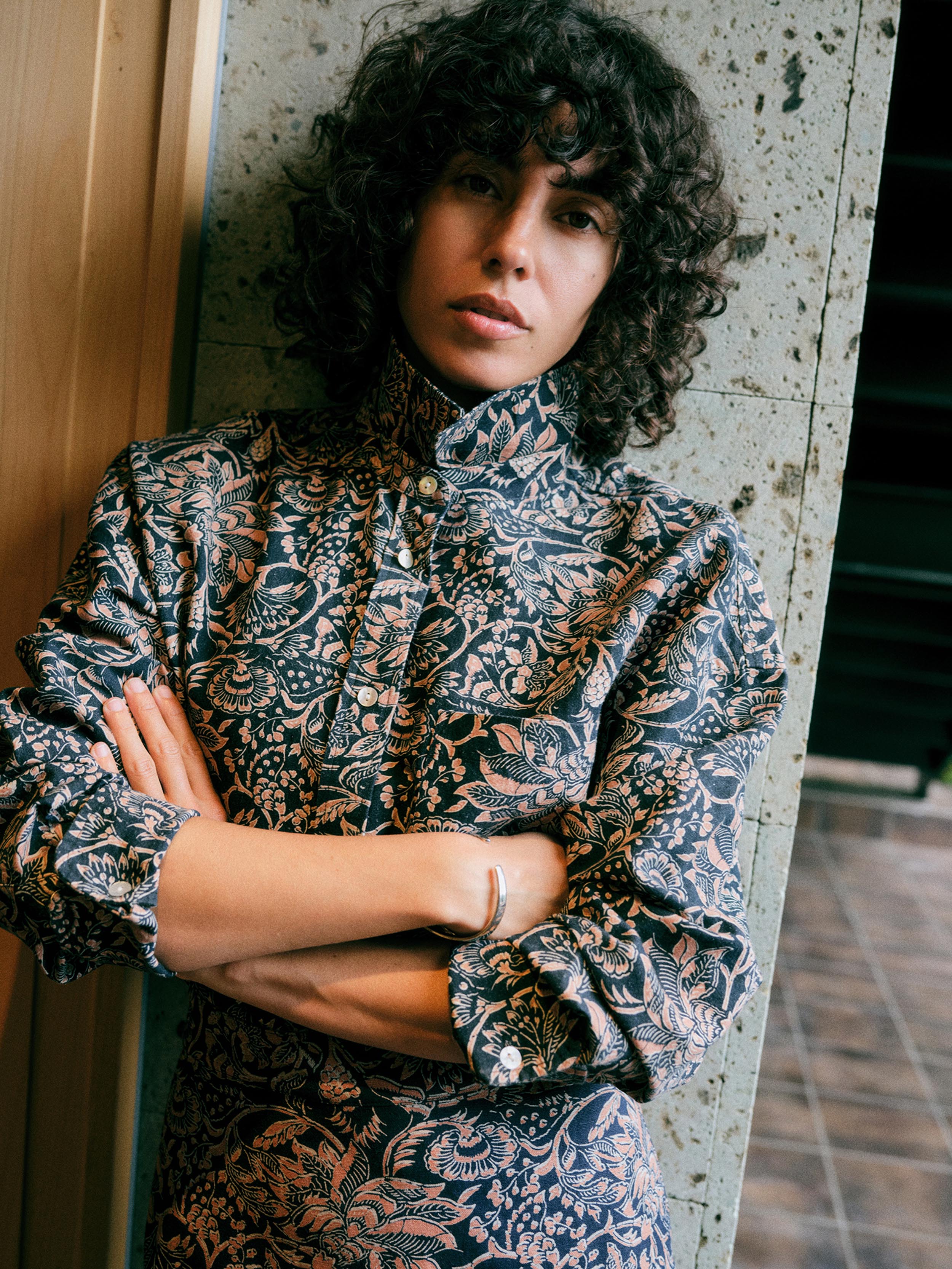 A woman with curly dark hair wearing a high-neck, long-sleeve button-up shirt with a dark navy and beige botanical pattern, leaning against a textured concrete pillar.