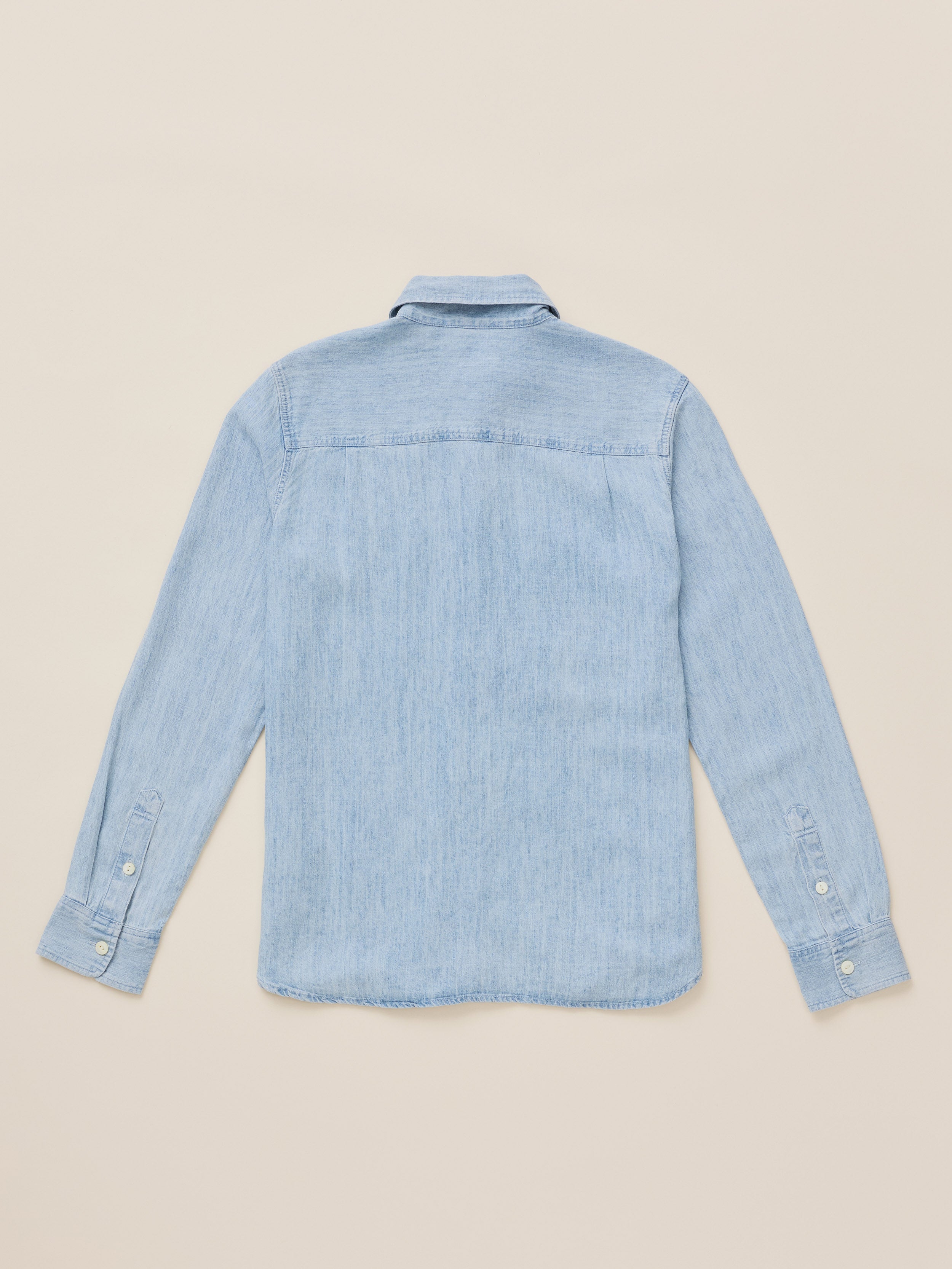 A light blue chambray long-sleeve button-up shirt shown from the back, laid flat on a warm beige background.