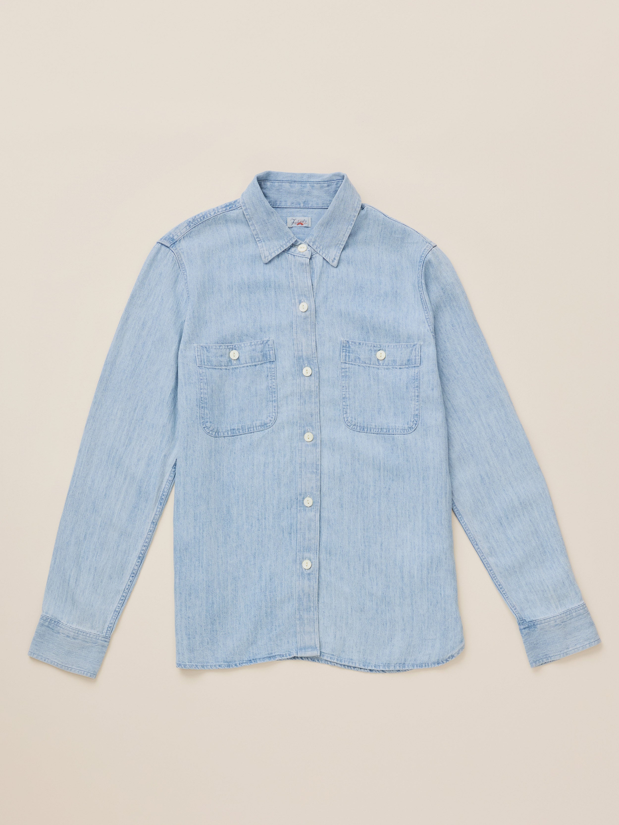 A light blue denim long-sleeve button-up shirt with two chest pockets, laid flat on a warm beige background.