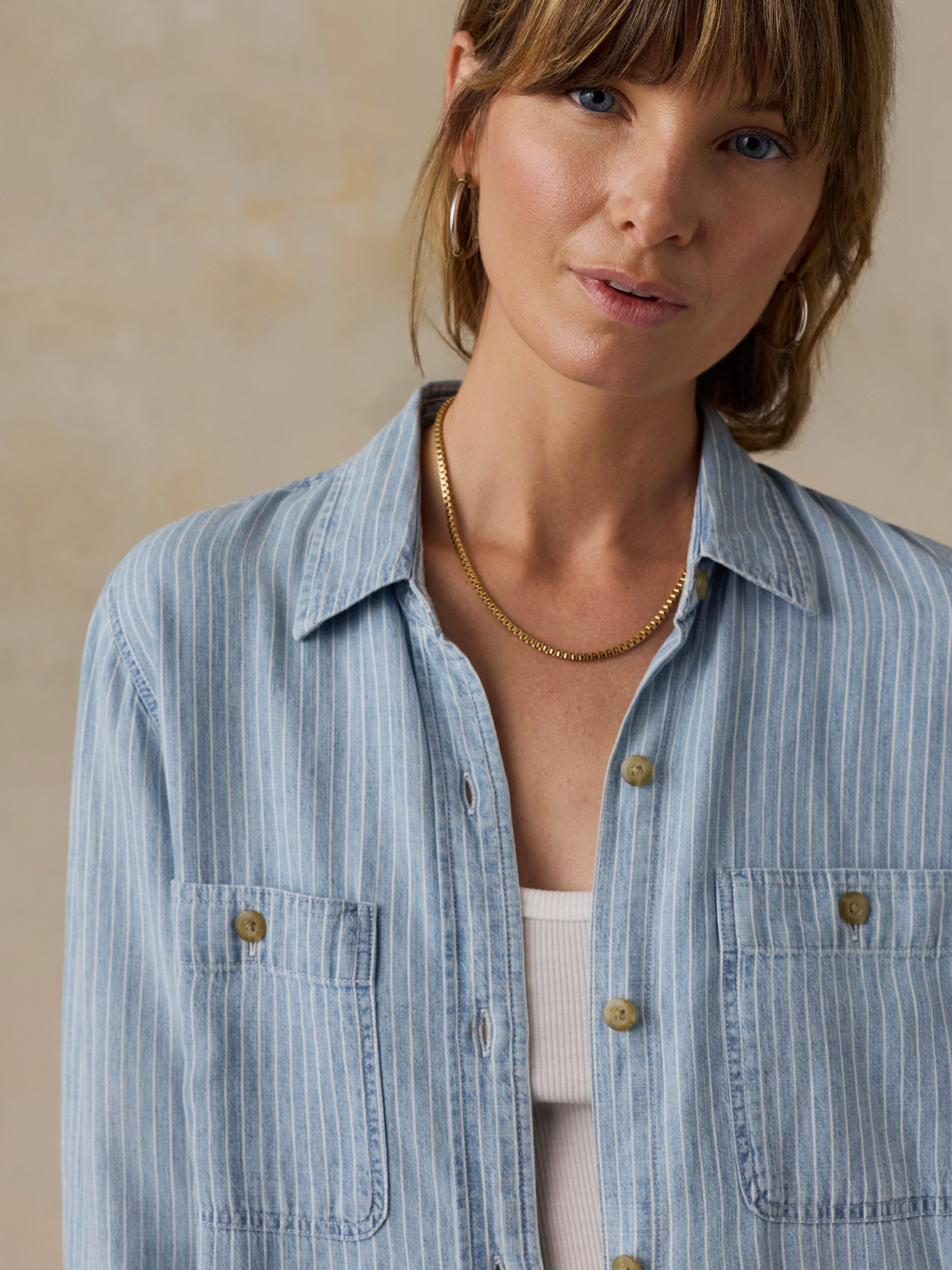 Woman wearing a blue and white striped chambray button down shirt over a white tank top against a beige background