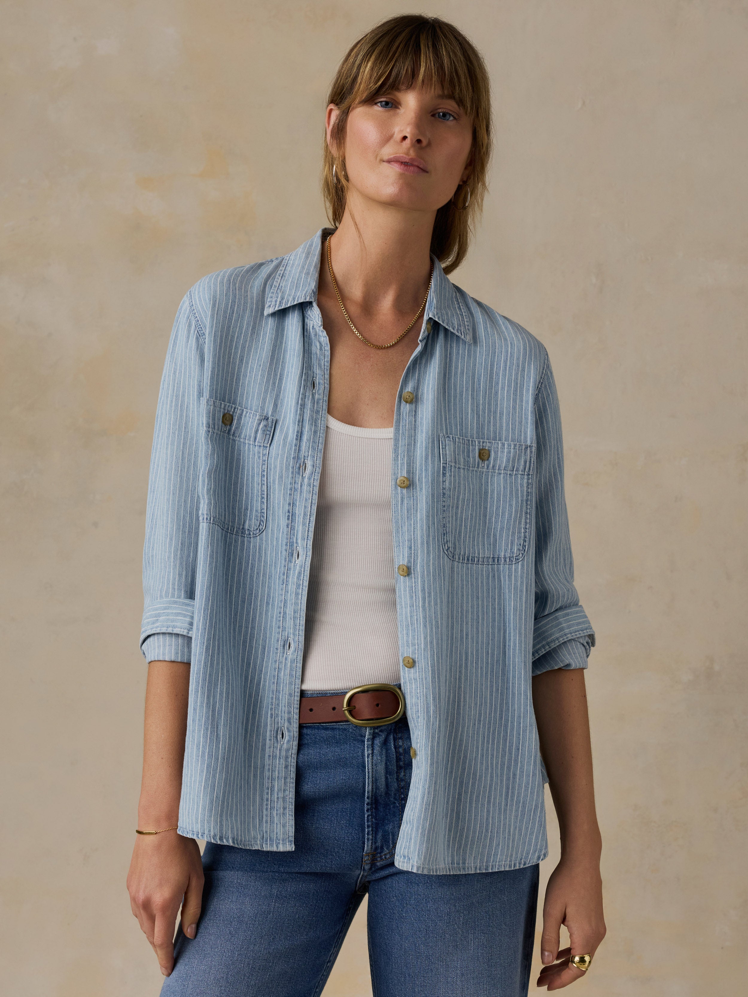 Woman wearing a blue and white striped chambray button down shirt over a white tank top and blue jeans against a beige background