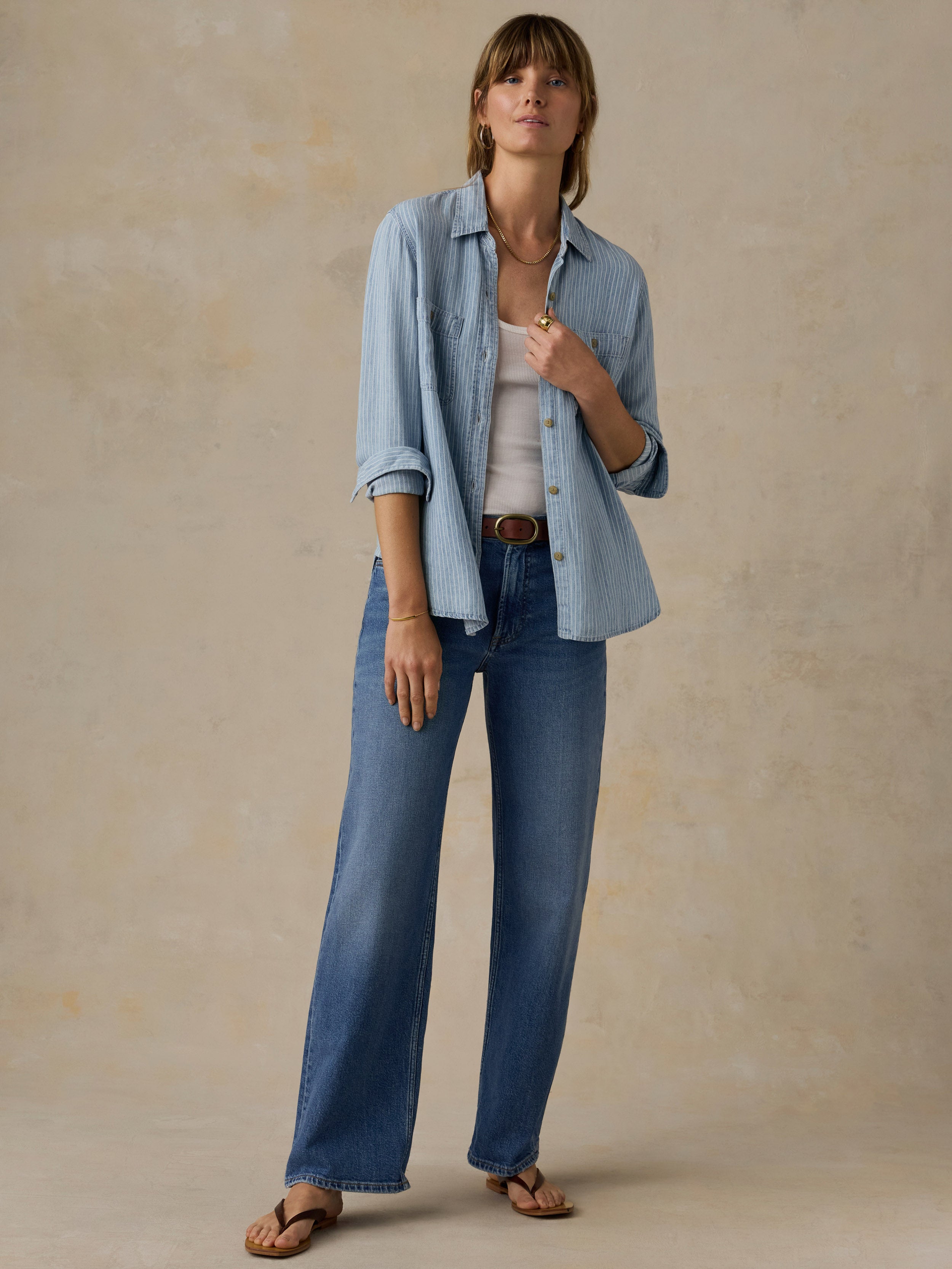 Woman wearing a blue and white striped chambray button down shirt over a white tank top and blue jeans against a beige background