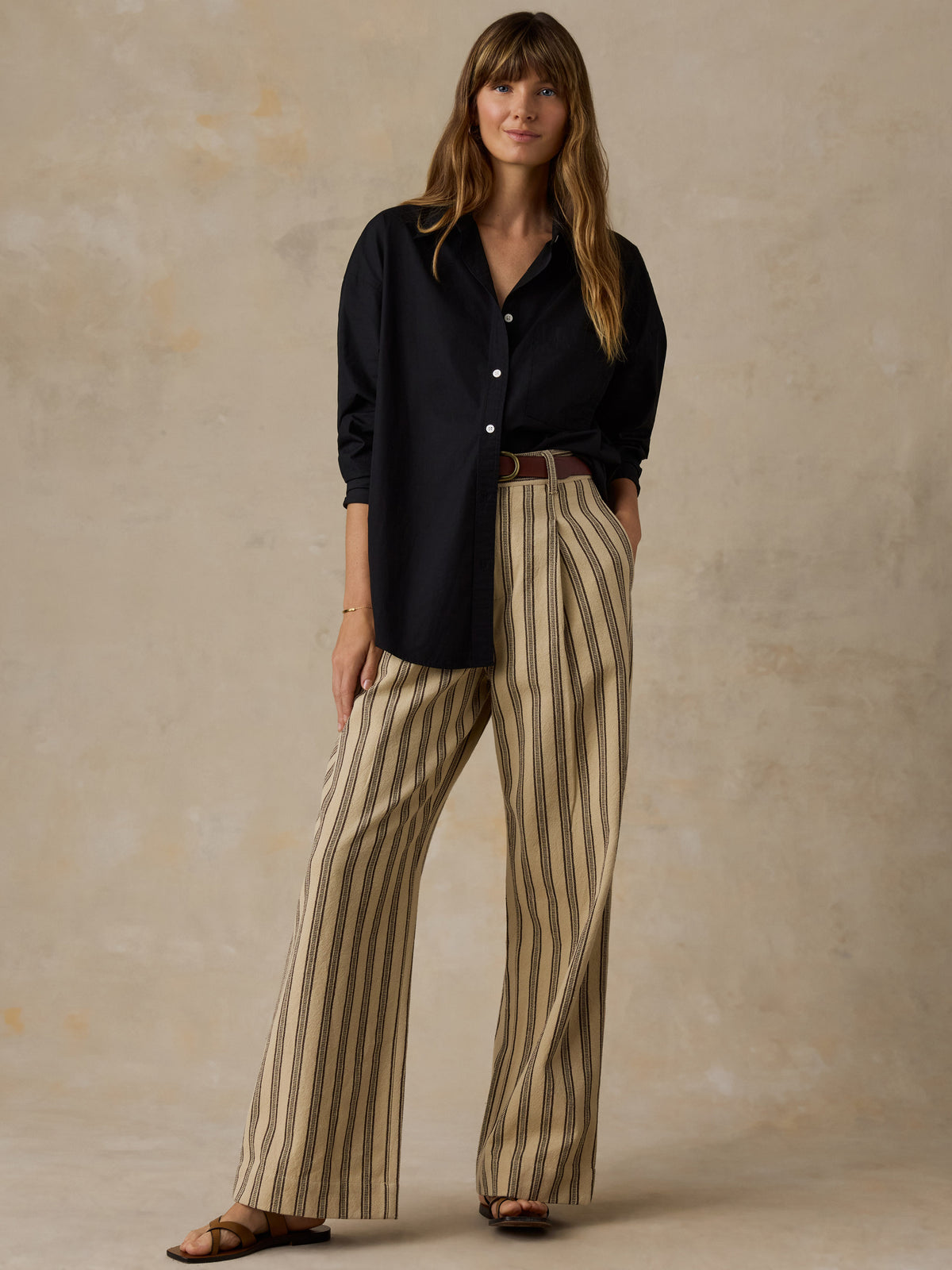 Woman wearing a black button down shirt and beige striped pants against a beige background, image 4 of 6