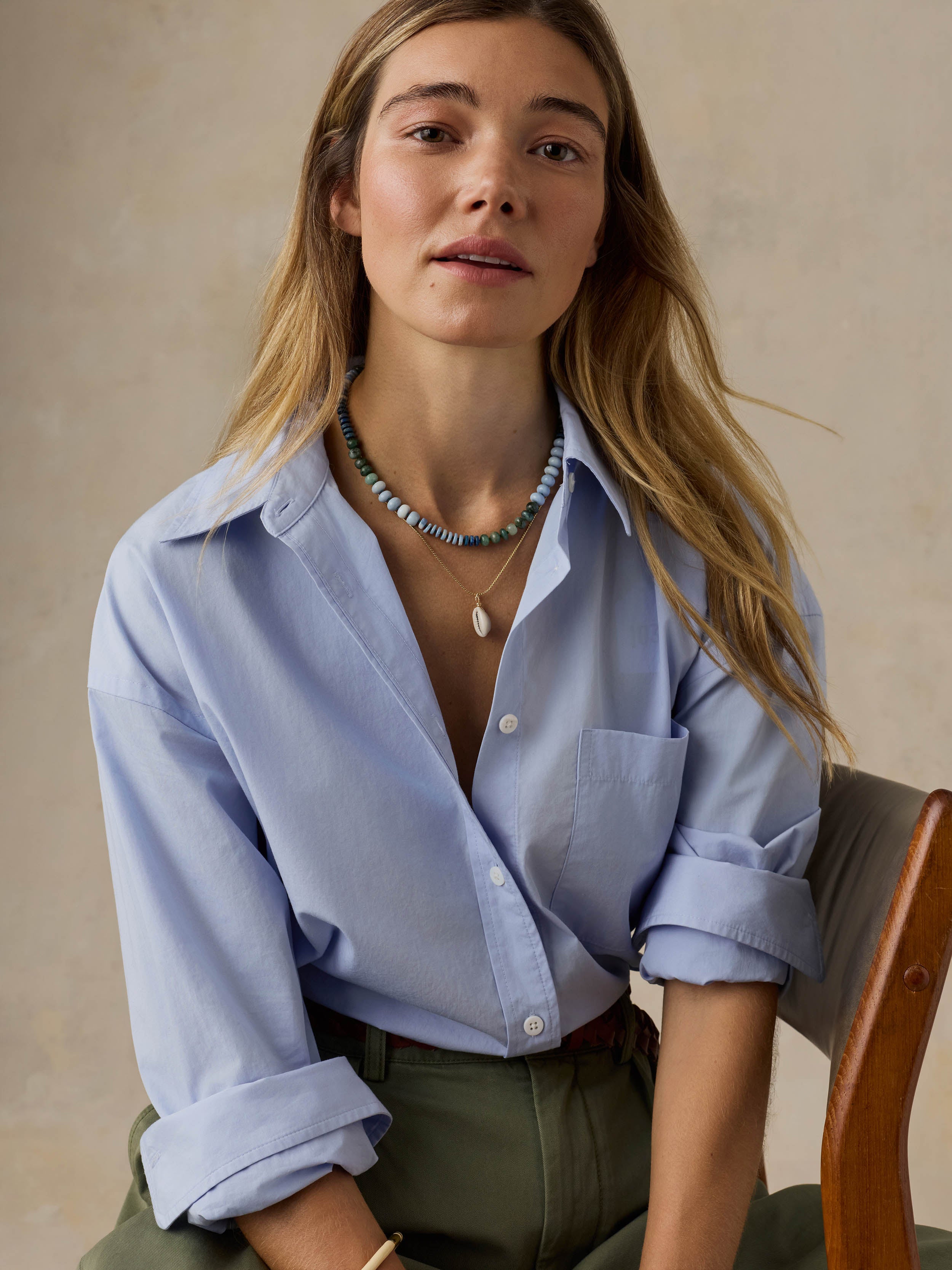 Woman sitting on a wooden chair wearing a light blue shirt and green pants against a beige background