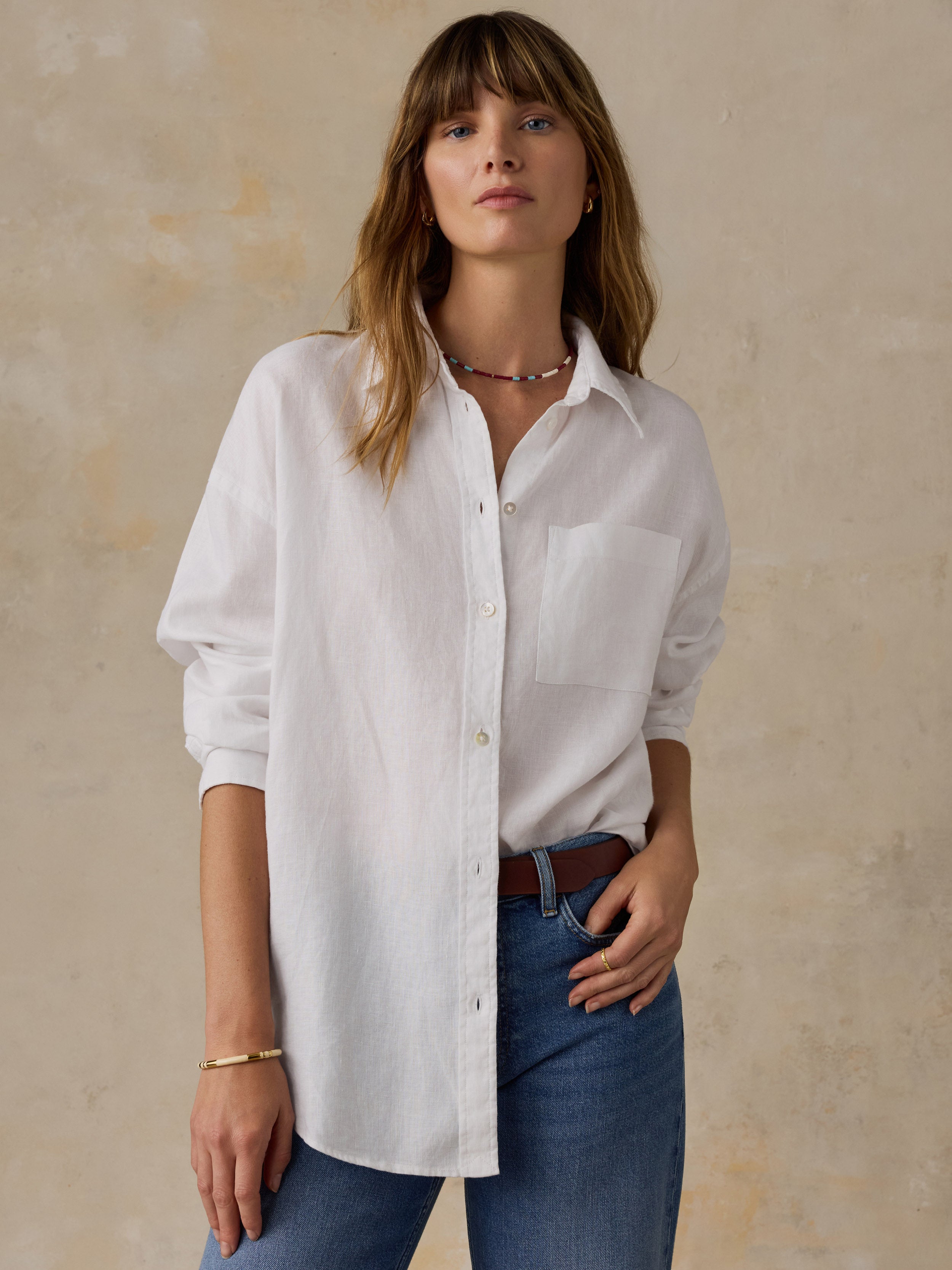 Woman wearing a white linen button down shirt with blue jeans against a beige background