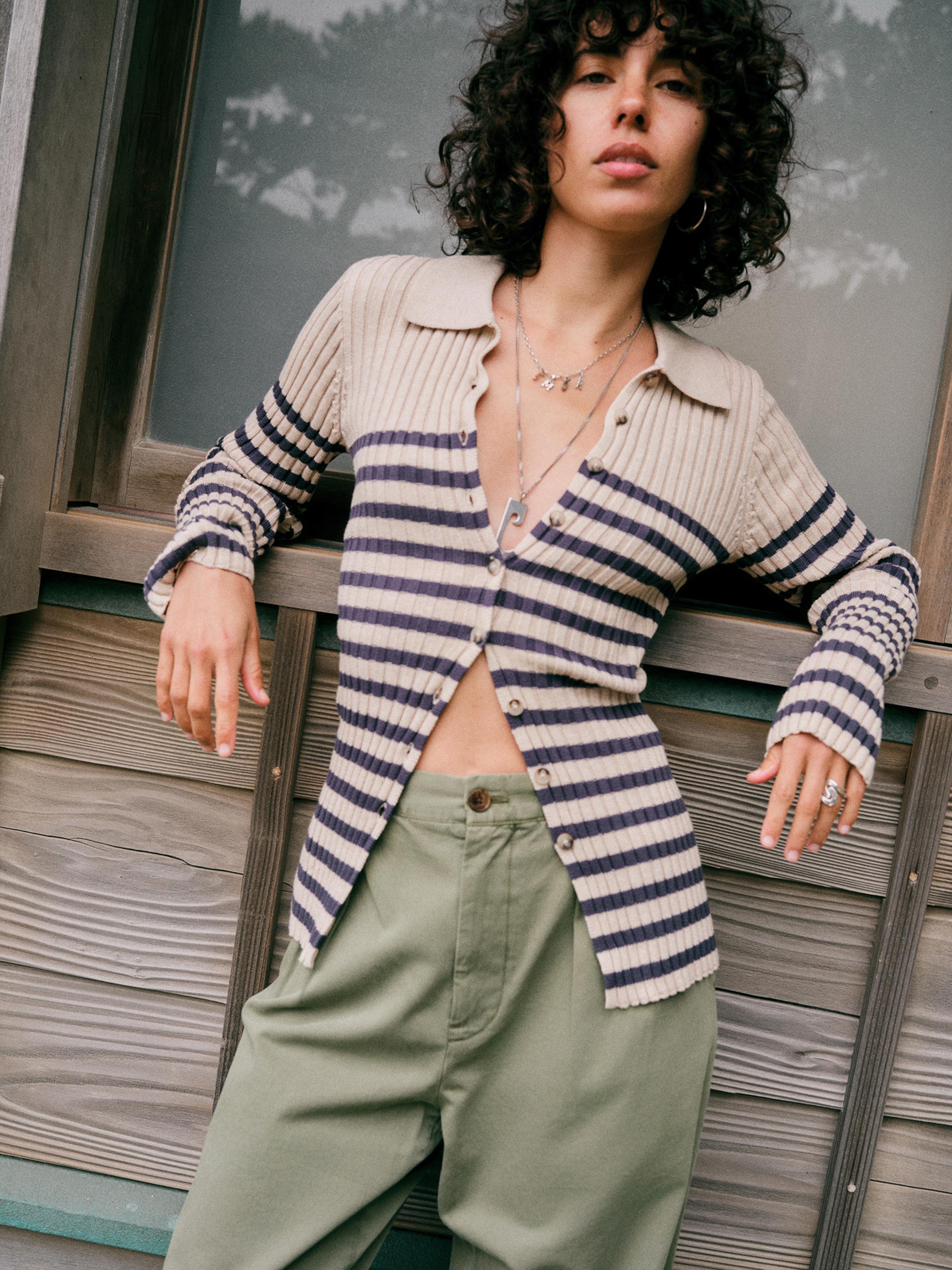 A model with curly black hair leans against a rustic wooden wall, wearing a beige and navy blue horizontally striped ribbed knit cardigan and sage green pants.