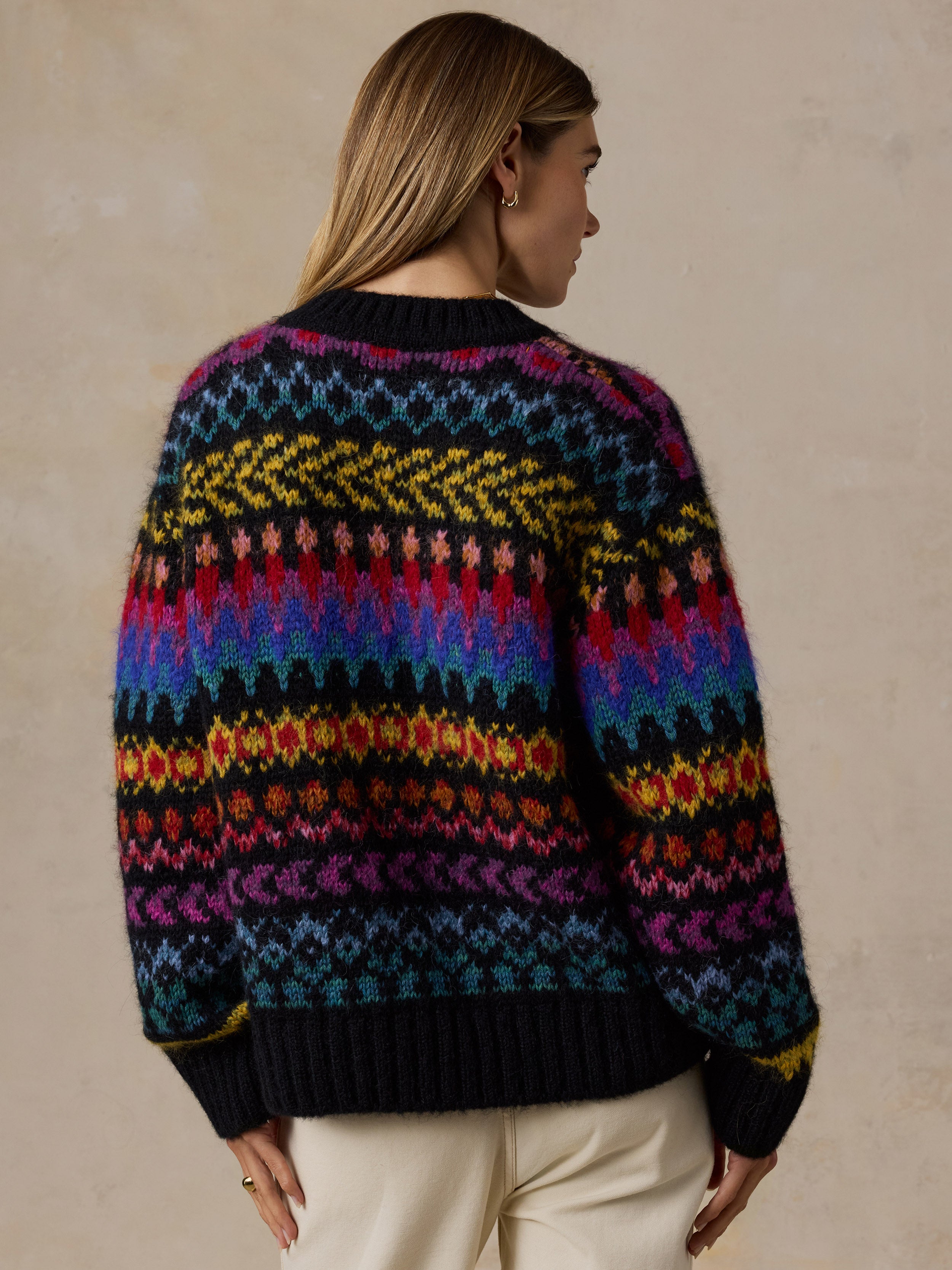 A woman viewed from the back wearing a colorful Fair Isle knit sweater with horizontal bands of blue, purple, pink, yellow, orange, and black, paired with cream pants against a textured beige wall., image 4 of 7