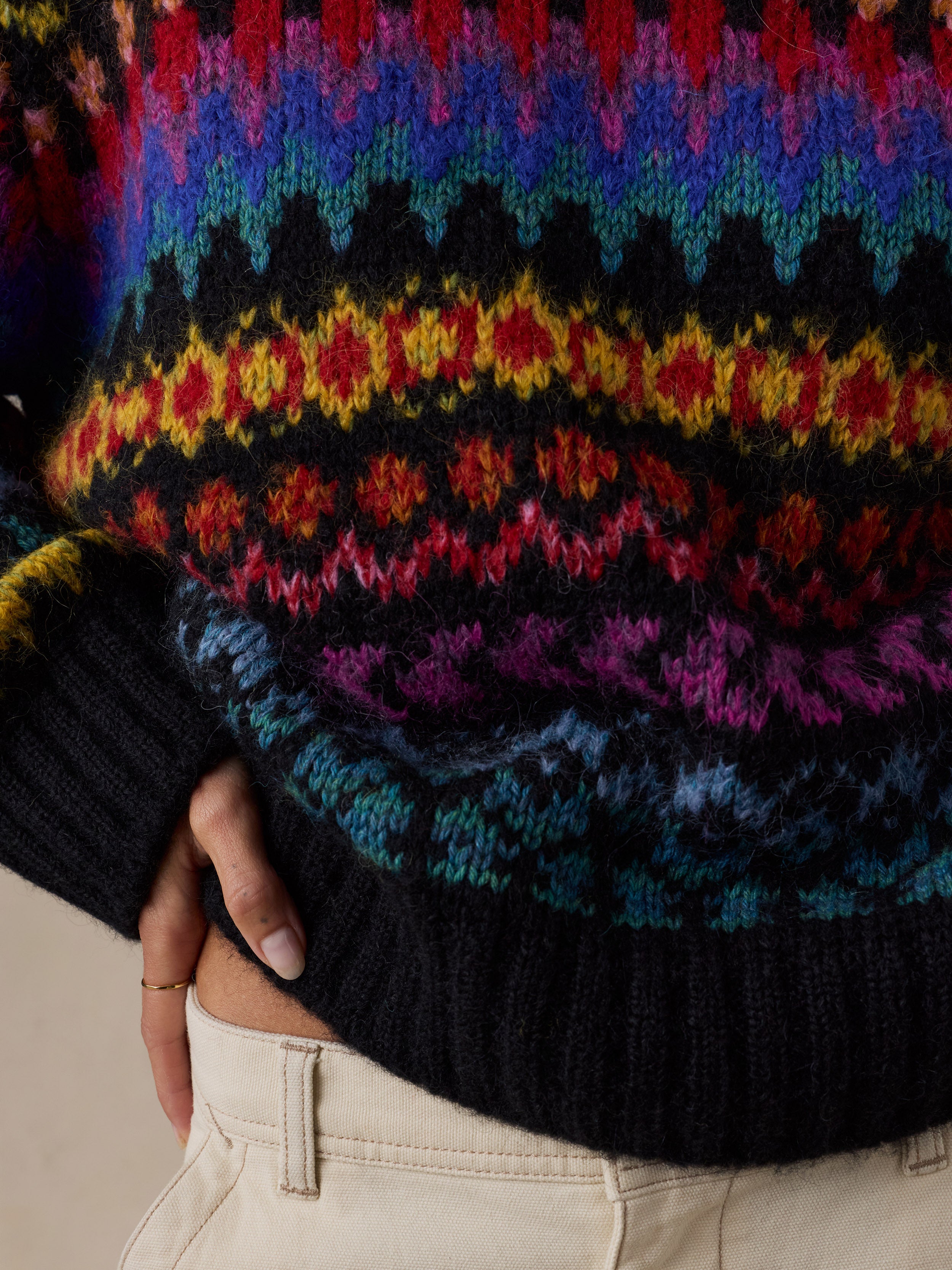 Close-up of a woman wearing a colorful Fair Isle knit sweater with a black ribbed hem and cuffs, paired with light beige pants., image 5 of 7