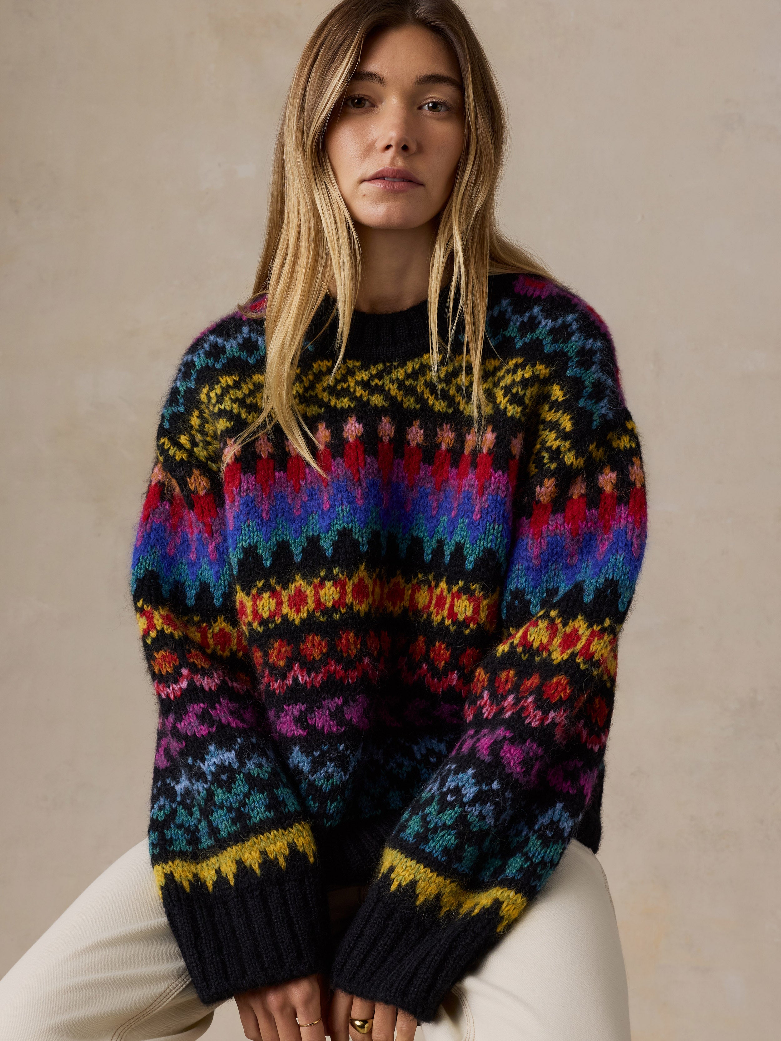 A woman wearing a colorful, patterned knit sweater with horizontal bands of yellow, red, blue, purple, and black, paired with cream-colored pants, posed against a neutral beige background., image 3 of 7