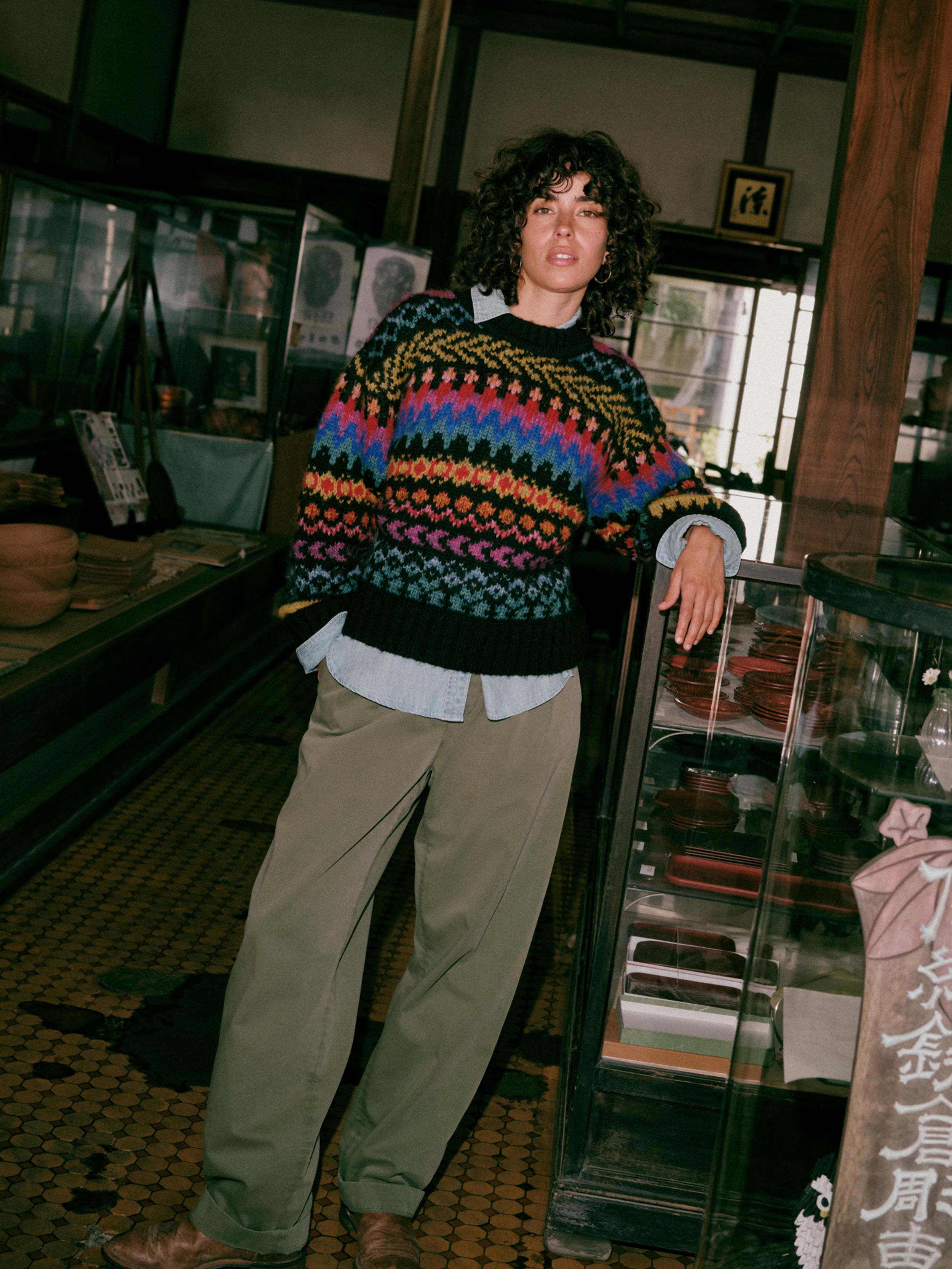 A woman standing in a shop interior wearing a colorful patterned knit sweater over a light blue shirt, with loose olive pants and brown shoes., image 1 of 7