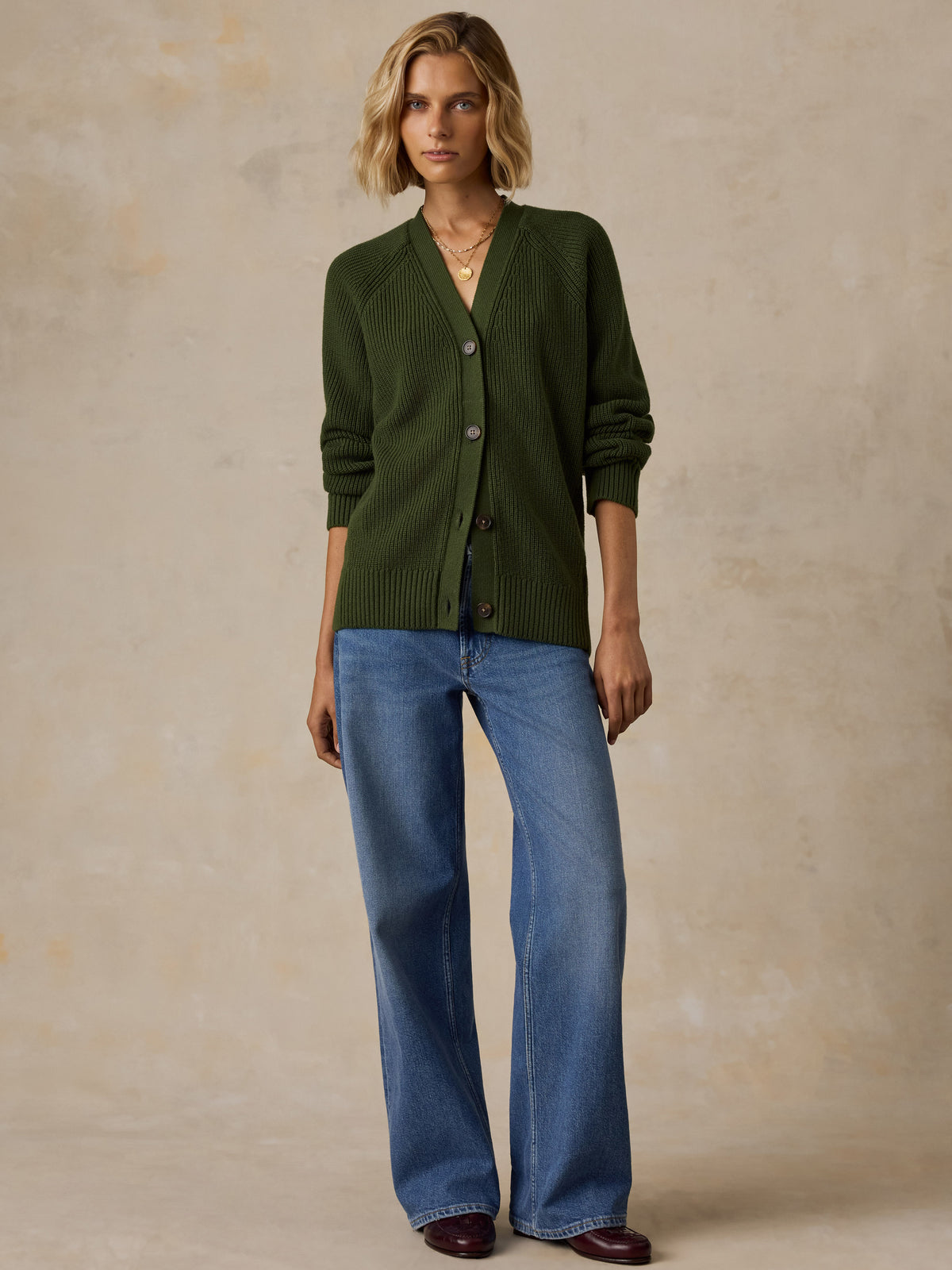 A woman wearing a dark olive green ribbed cardigan sweater with brown buttons, paired with blue jeans and dark brown shoes, image 6 of 6