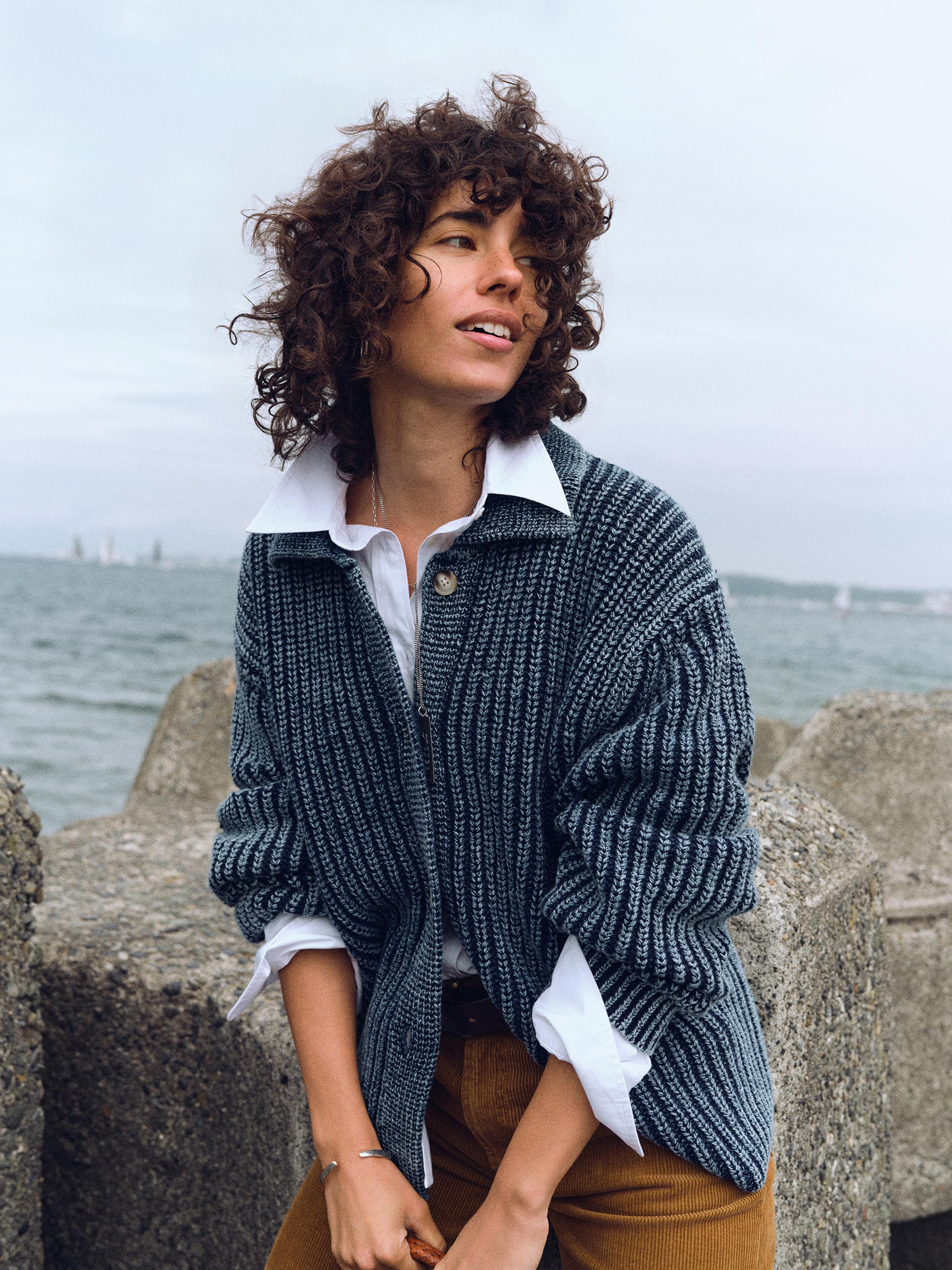 A woman with curly brown hair sits outdoors on stone breakwaters with the ocean in the background, wearing a dark blue-gray ribbed knit cardigan over a white collared shirt and brown pants.