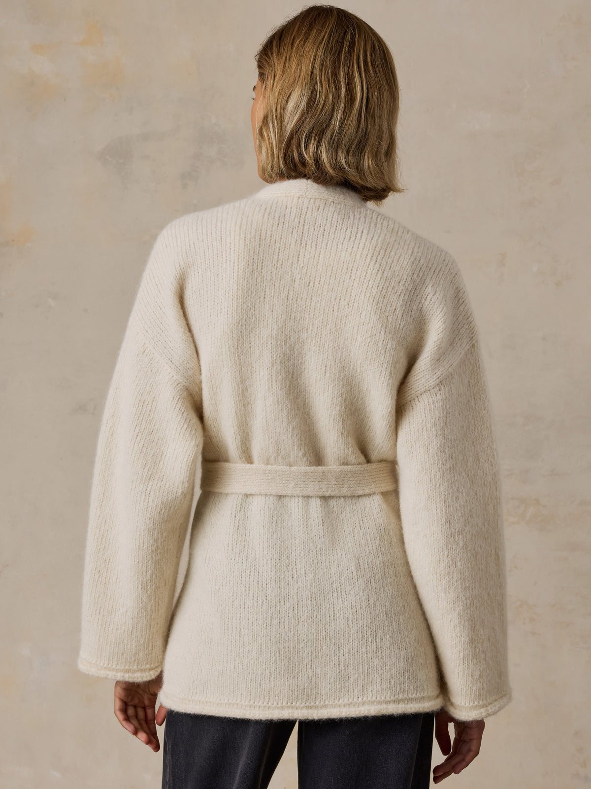 A woman viewed from the back wearing a cream wrap cardigan and black jeans, image 2 of 8