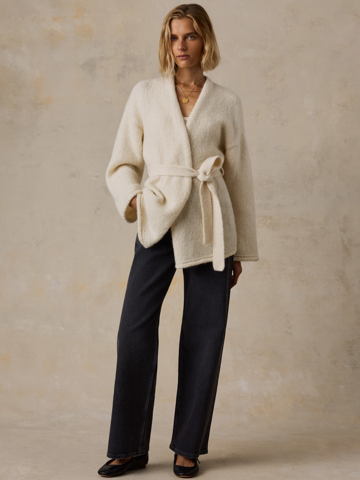 A woman wearing a cream wrap cardigan, black jeans, and black flats, image 7 of 8