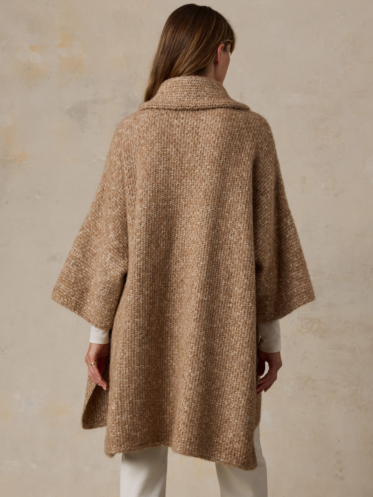 A woman viewed from behind wearing a beige sweater poncho over a white turtleneck and white pants, image 4 of 7