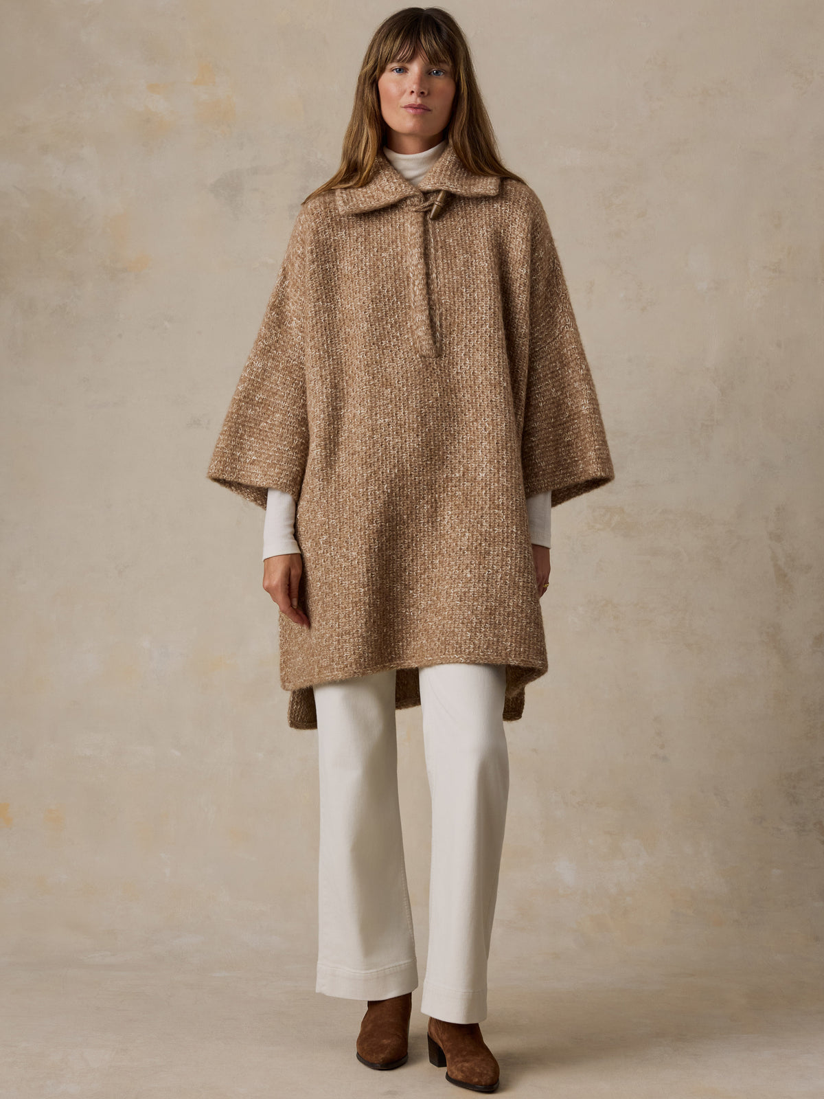 A woman wearing a beige sweater poncho with a white turtleneck, white pants, and suede boots, image 7 of 7