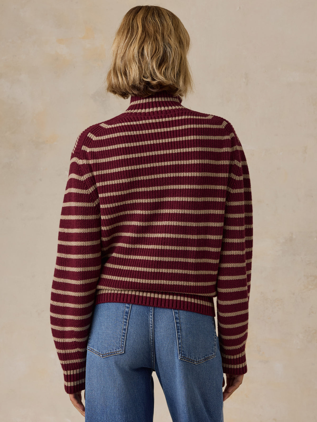A woman wearing a burgundy and beige striped quarter-zip knit sweater with blue jeans, image 3 of 5